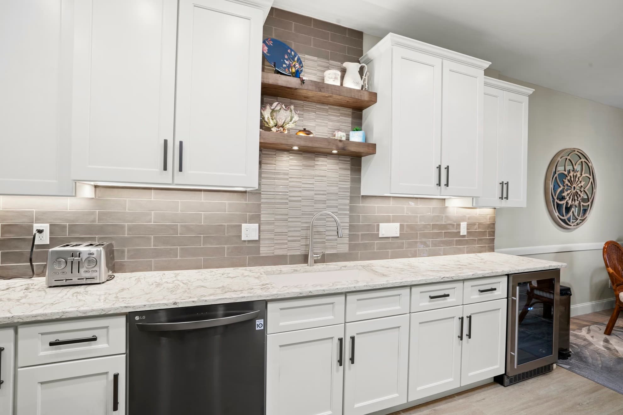 White shaker kitchen cabinets with two wood floating shelves over a grey tile backsplash with a mosaic accent, and a white veined countertop with an undermount sink