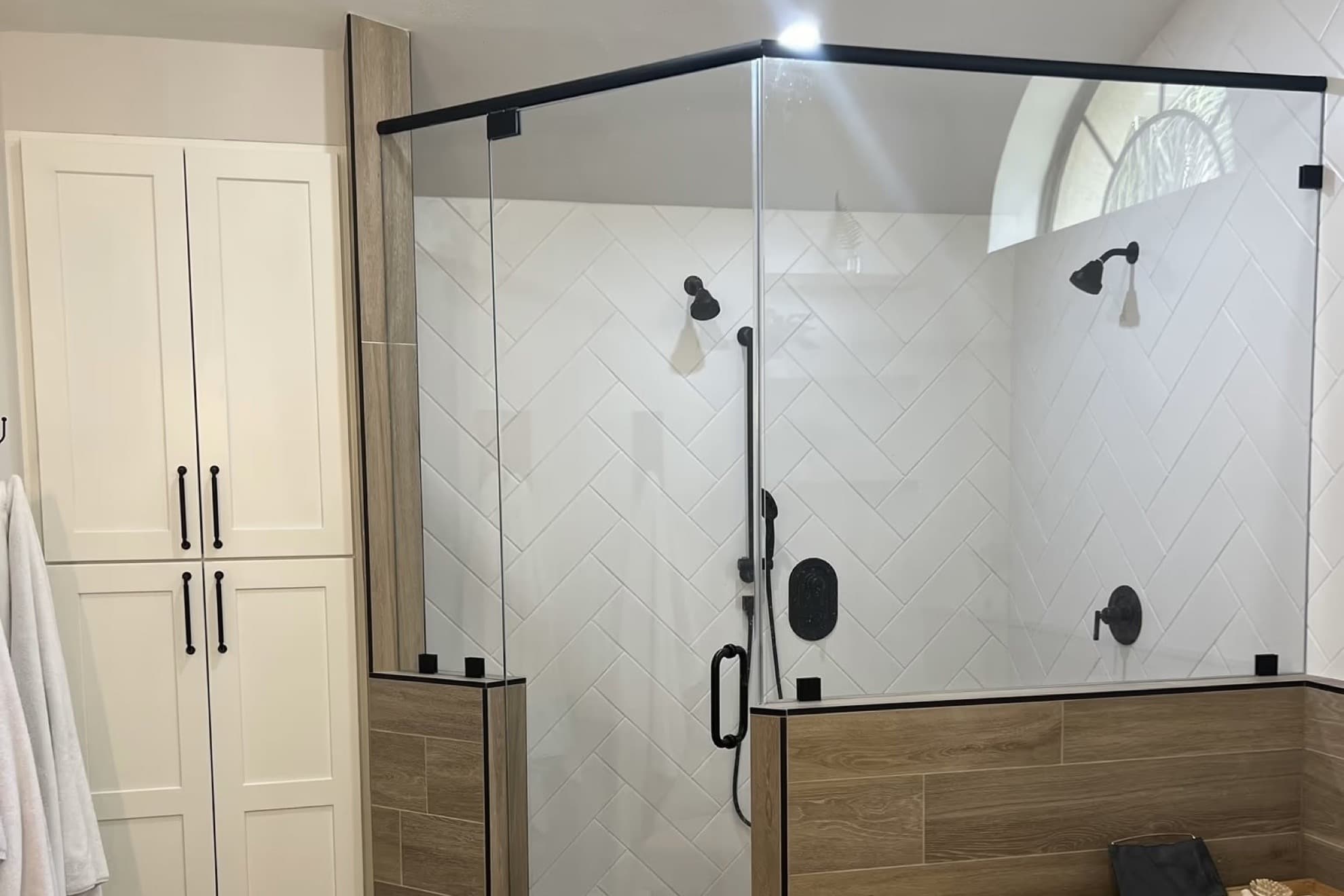 Angled glass shower enclosure with a black top bar and black clips on wood-look tile half walls, with white herringbone tile and two black shower heads inside