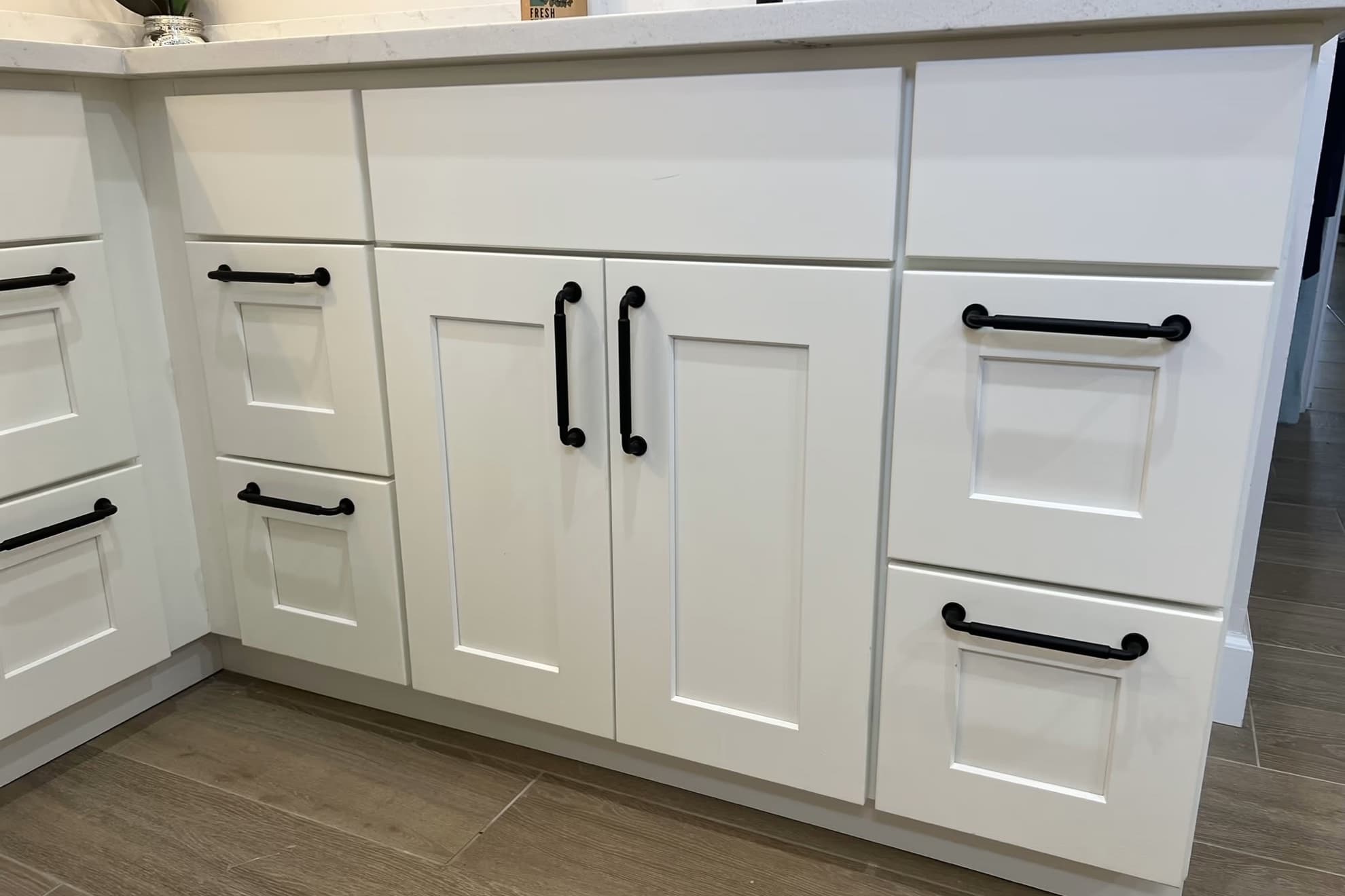 Close-up of white shaker vanity cabinets with black bar pulls beneath a white veined countertop, on wood-look tile flooring