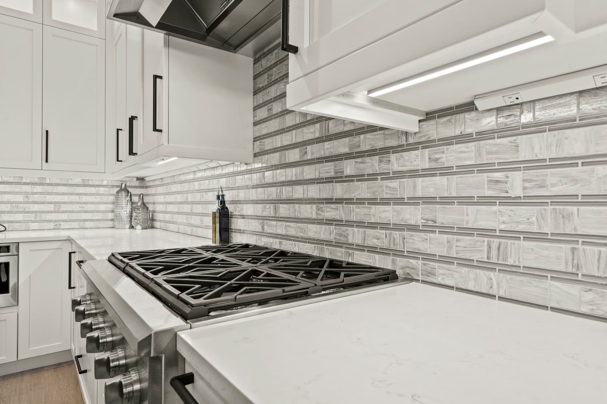 Narrow gray and white glass tile backsplash behind a stainless gas cooktop, under white cabinets with a black hood and an under-cabinet light strip