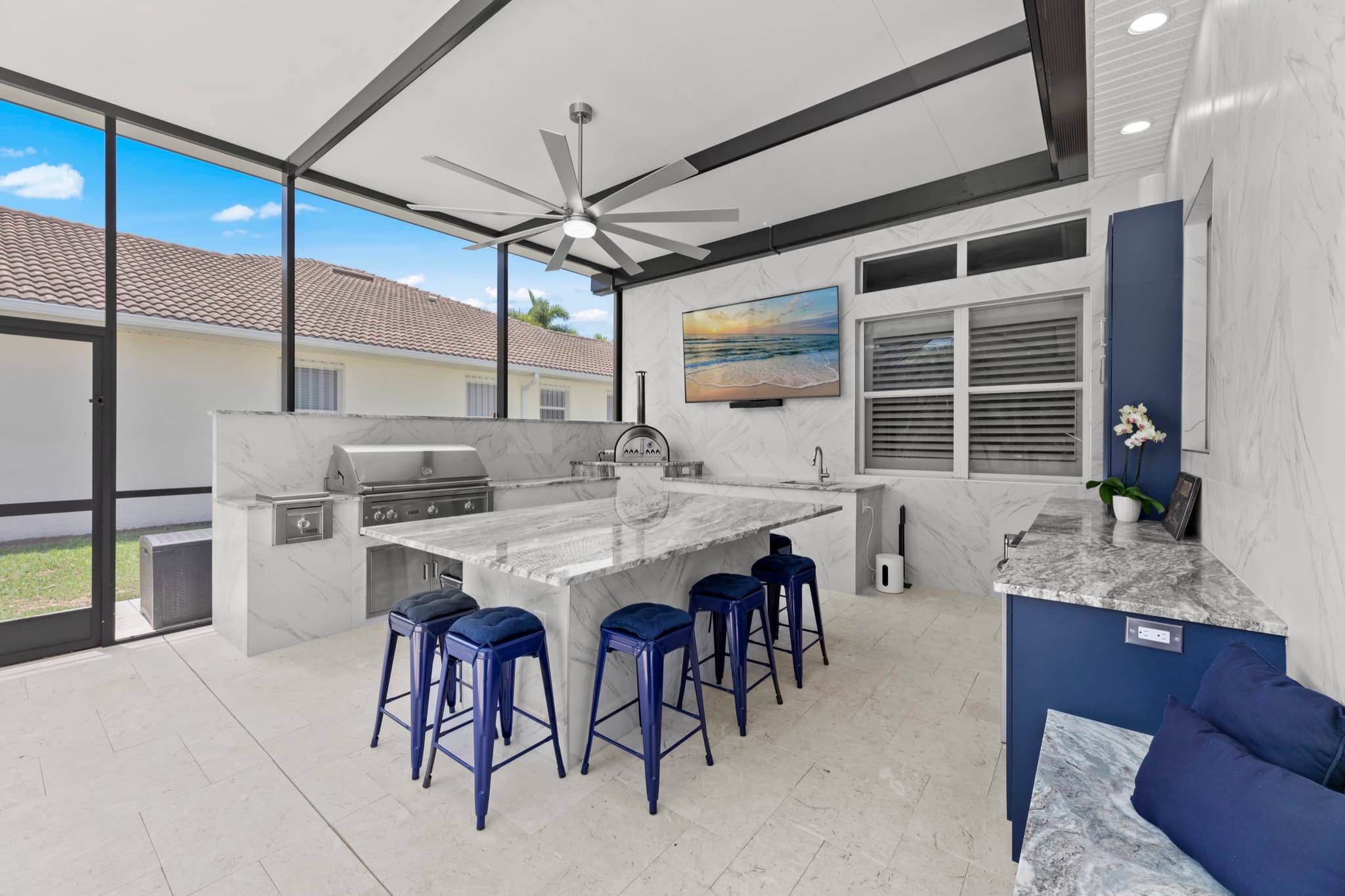 Screened lanai outdoor kitchen with a marble-look island, navy bar stools, a built-in grill run, a ceiling fan, and the yard beyond