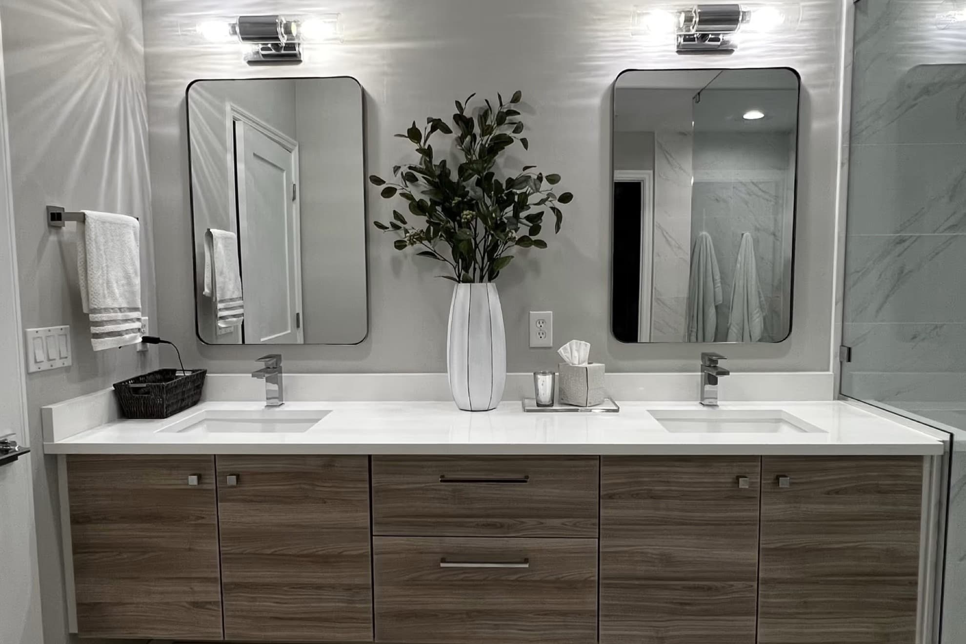 Remodeled bathroom with a wood-look double vanity, a white countertop with two undermount sinks, two framed mirrors and wall sconces, beside a glass shower