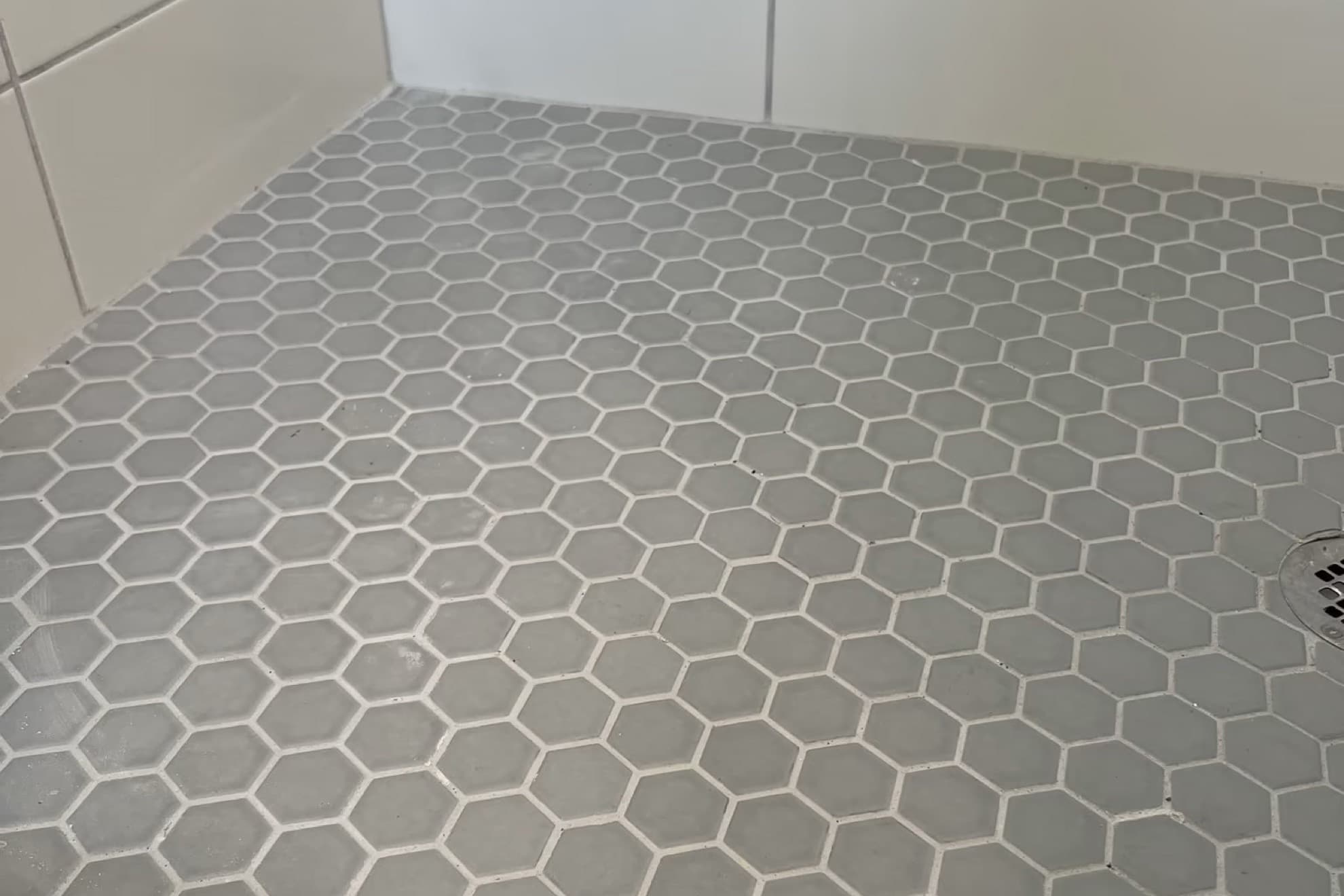 Grey hexagon mosaic tile shower floor meeting white rectangular wall tile, with the edge of a round drain at the right