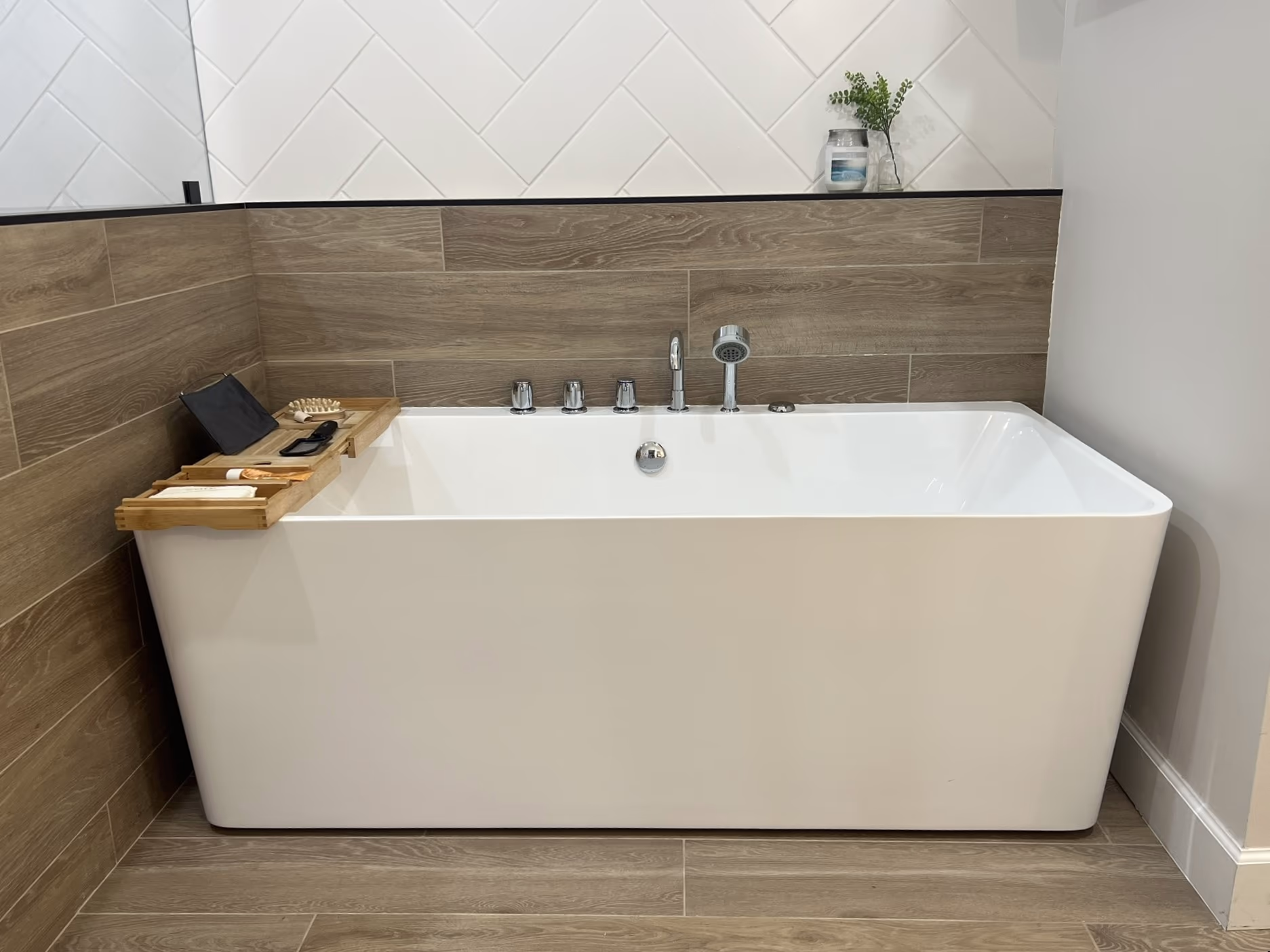 Freestanding white soaking tub with chrome fixtures, a wood bath caddy, and a wood-look tile surround