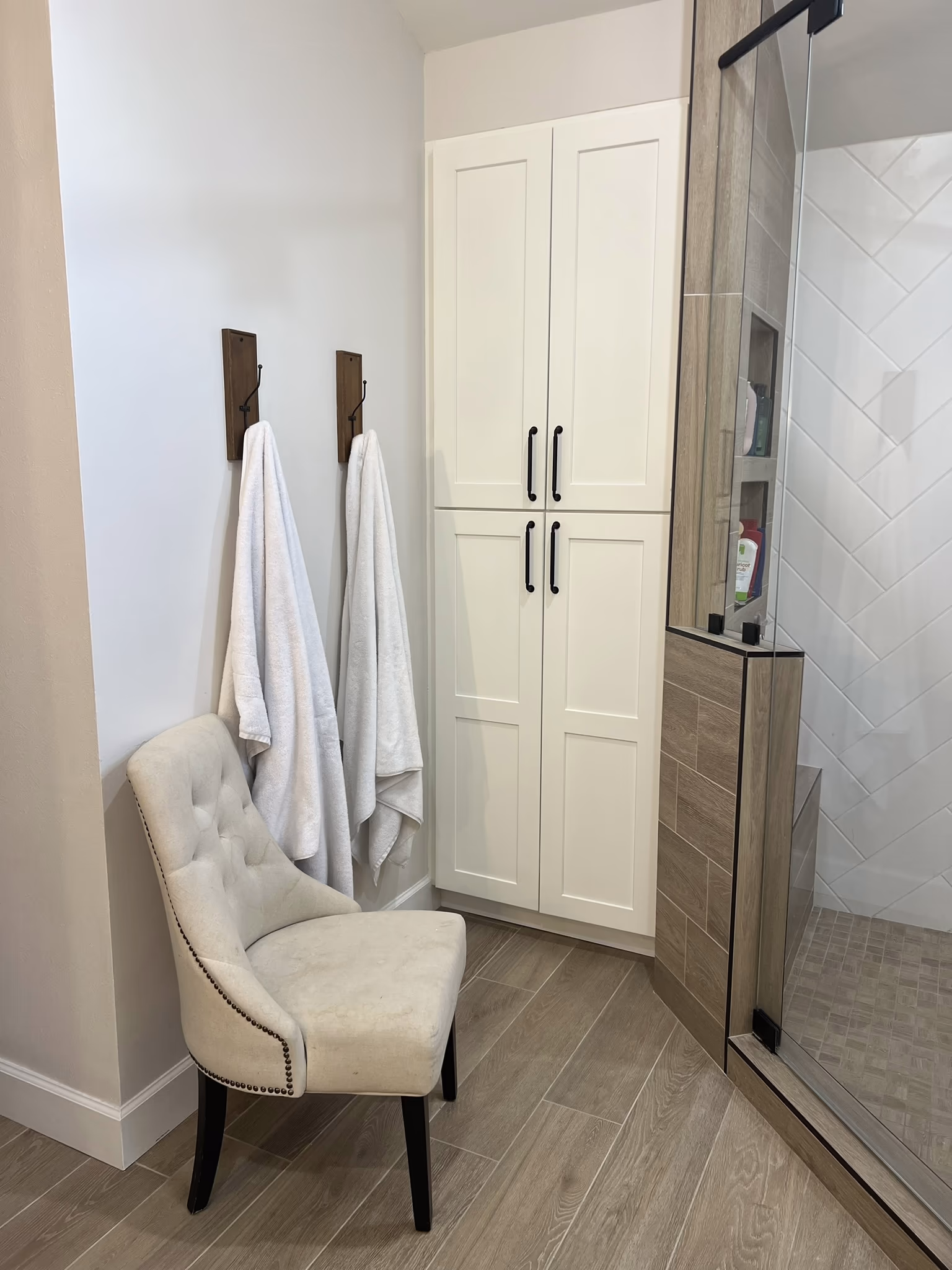 White linen cabinet in the corner of the bathroom, with towels on hooks and a tufted chair beside the shower door