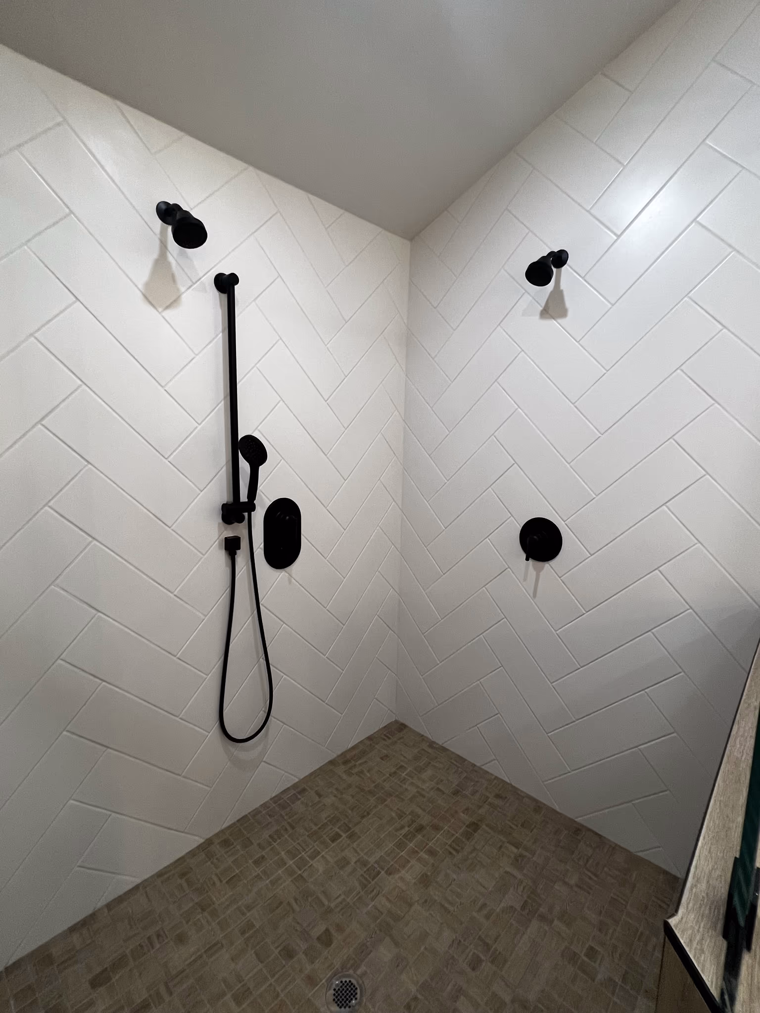 Inside a herringbone-tiled shower with two black shower heads and a mosaic tile floor