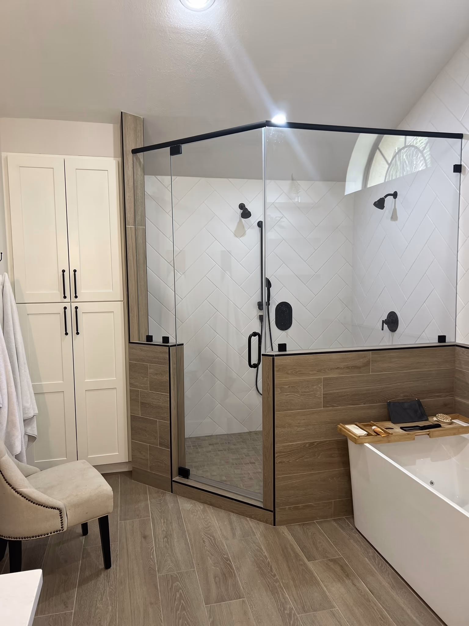 Corner glass shower exterior with a wood-tile half-wall, a tall white linen cabinet, and a tufted chair with towels on hooks