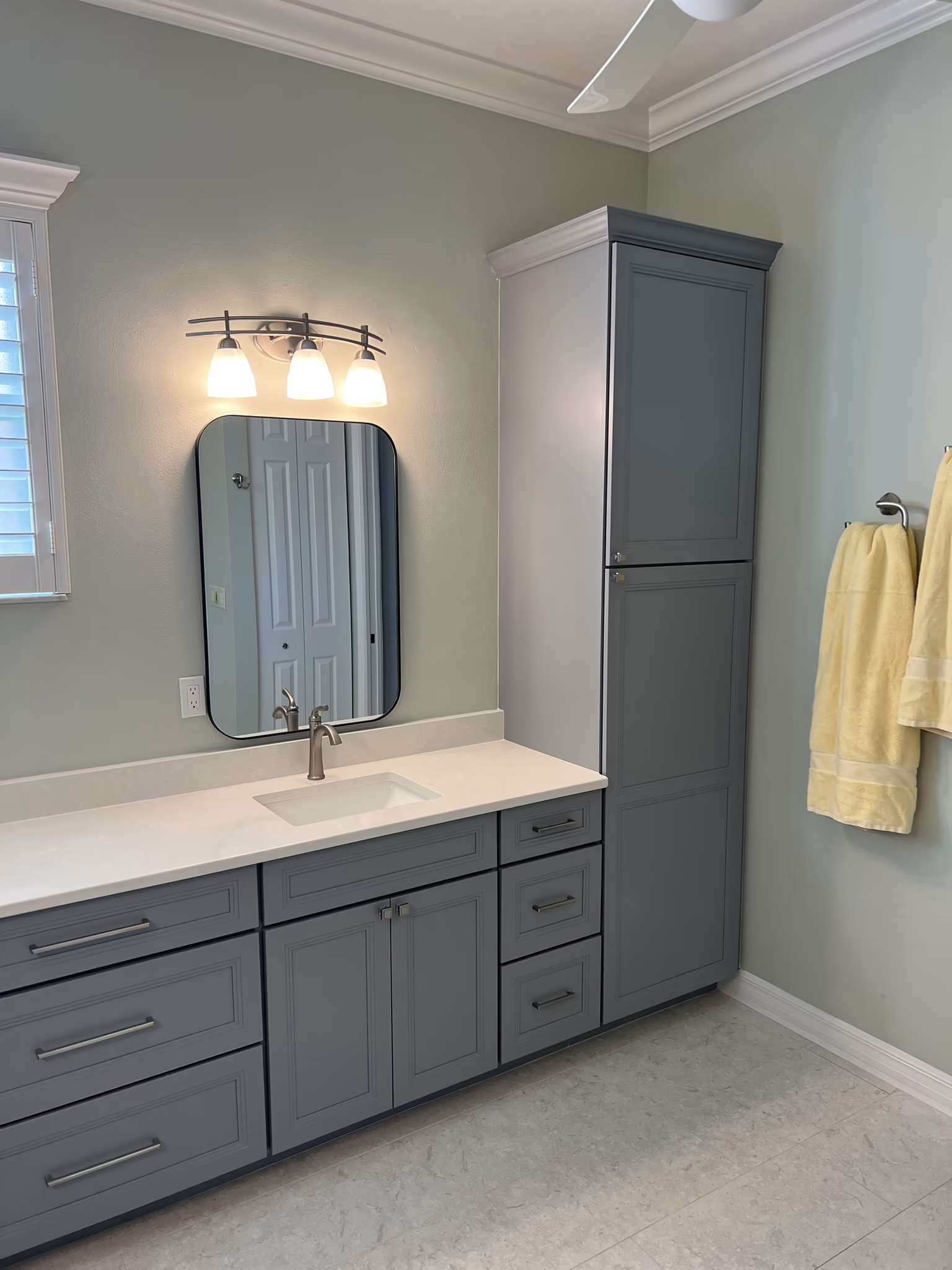 Grey shaker vanity and linen tower with a black oval mirror, three-light sconce, and yellow towels on hooks