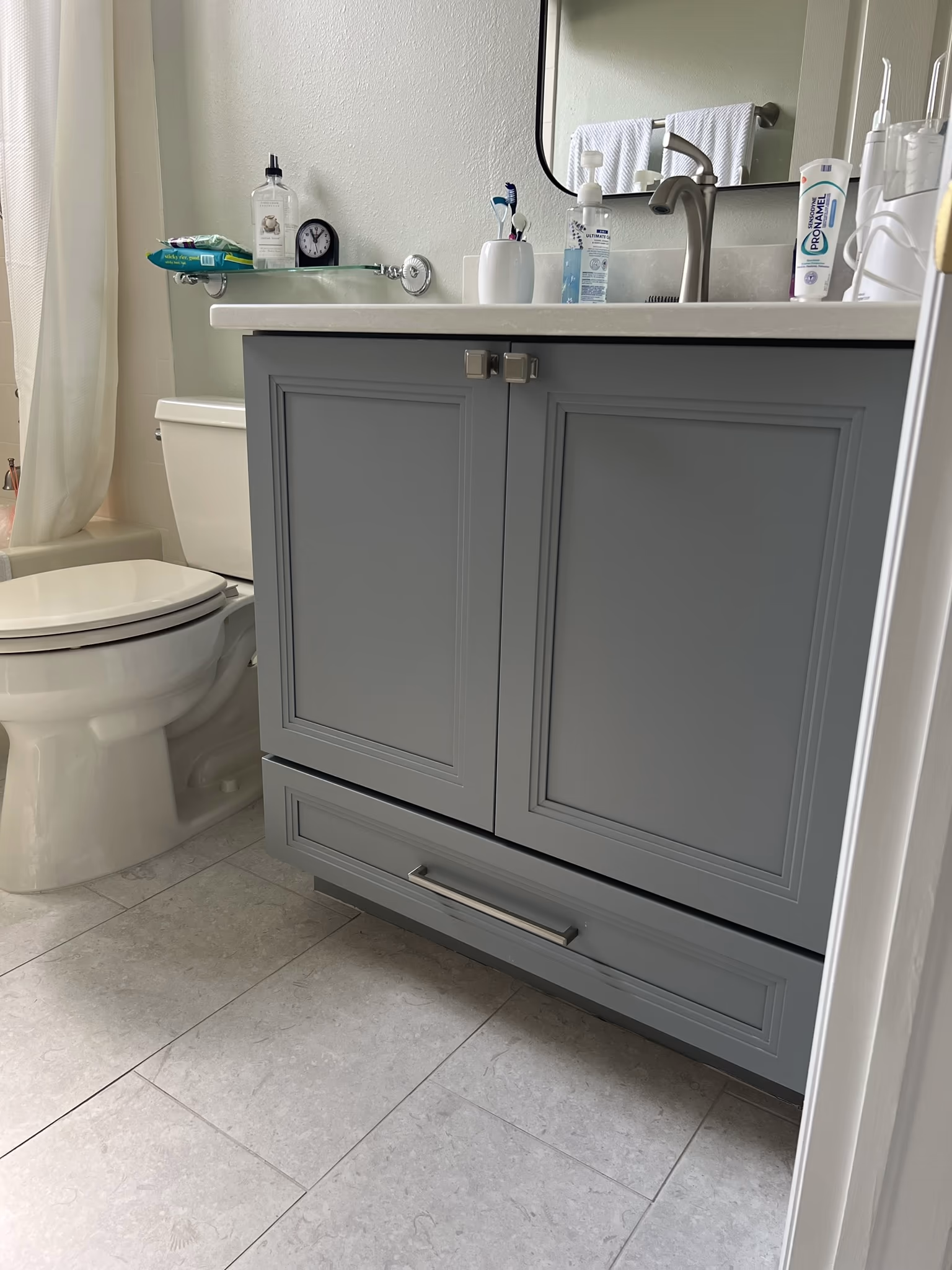 White toilet beside a grey vanity cabinet base on tile flooring, with a shower curtain visible at left