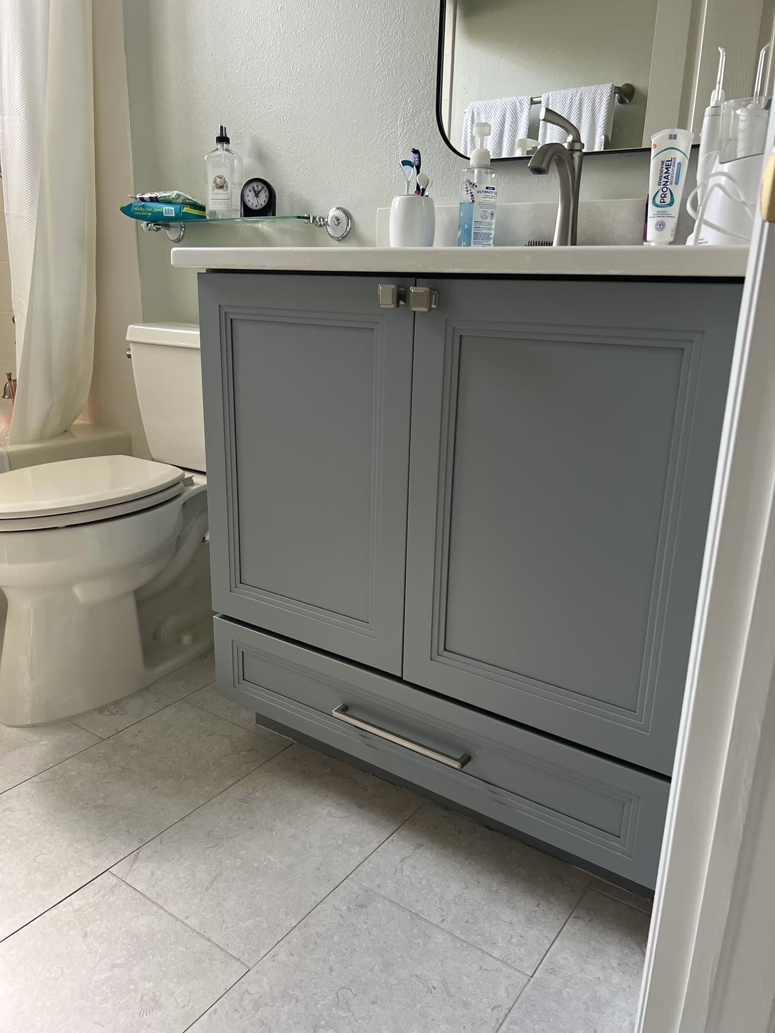 White toilet beside a grey vanity cabinet base on tile flooring, with a shower curtain visible at left