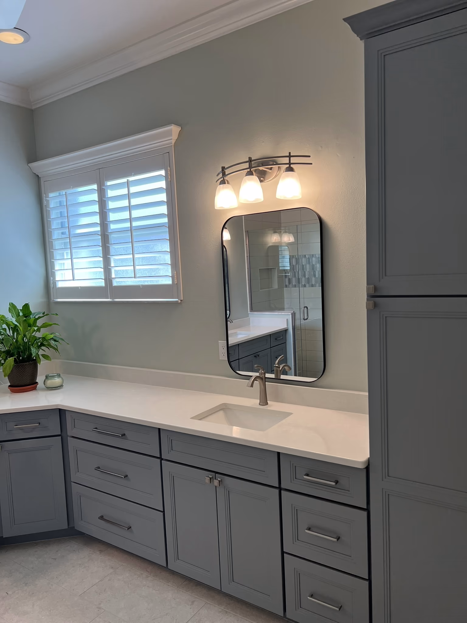 Bathroom vanity with grey shaker cabinets, a black oval mirror, three-light sconce, and a window with plantation shutters