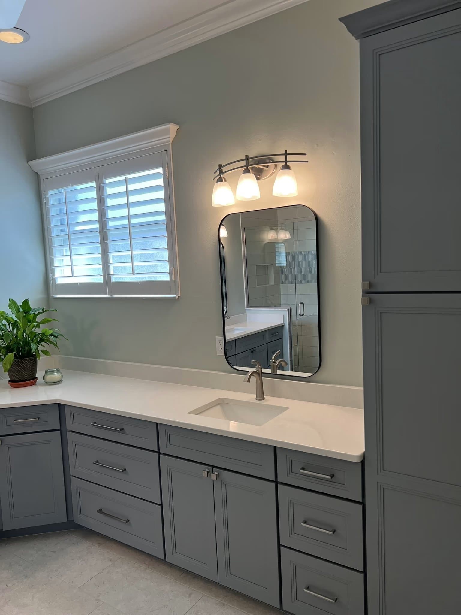 Bathroom vanity with grey shaker cabinets, a black oval mirror, three-light sconce, and a window with plantation shutters