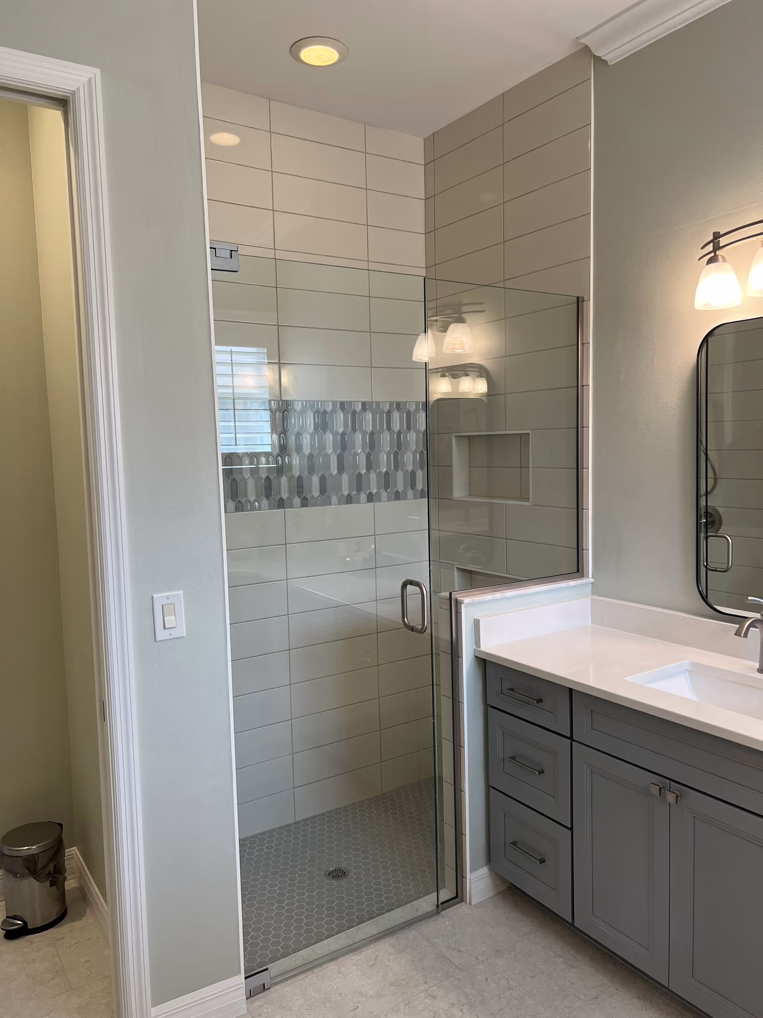 Glass shower door revealing tiled walls with a mosaic accent band, next to a vanity sink and cabinets