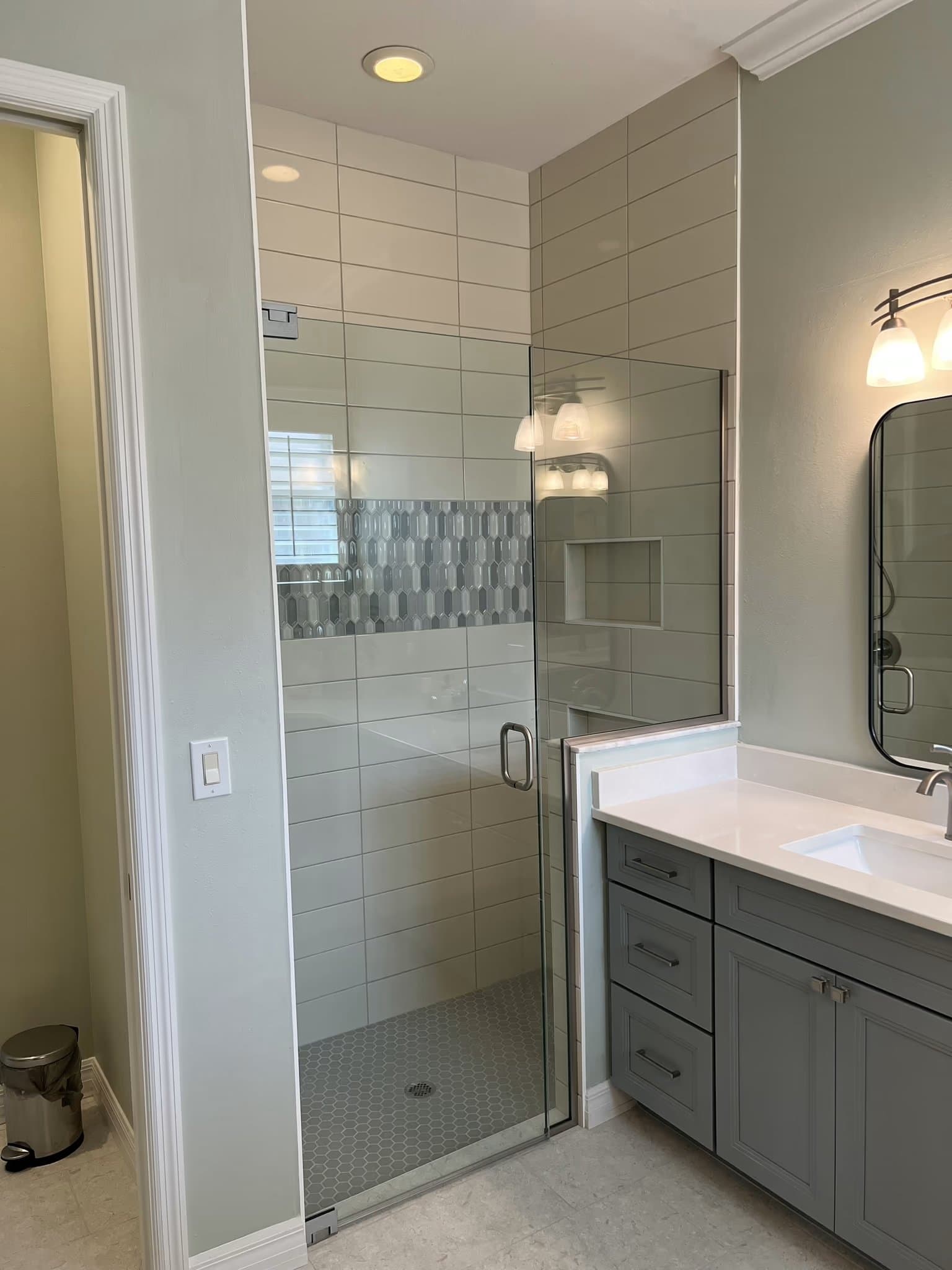 Glass shower door revealing tiled walls with a mosaic accent band, next to a vanity sink and cabinets