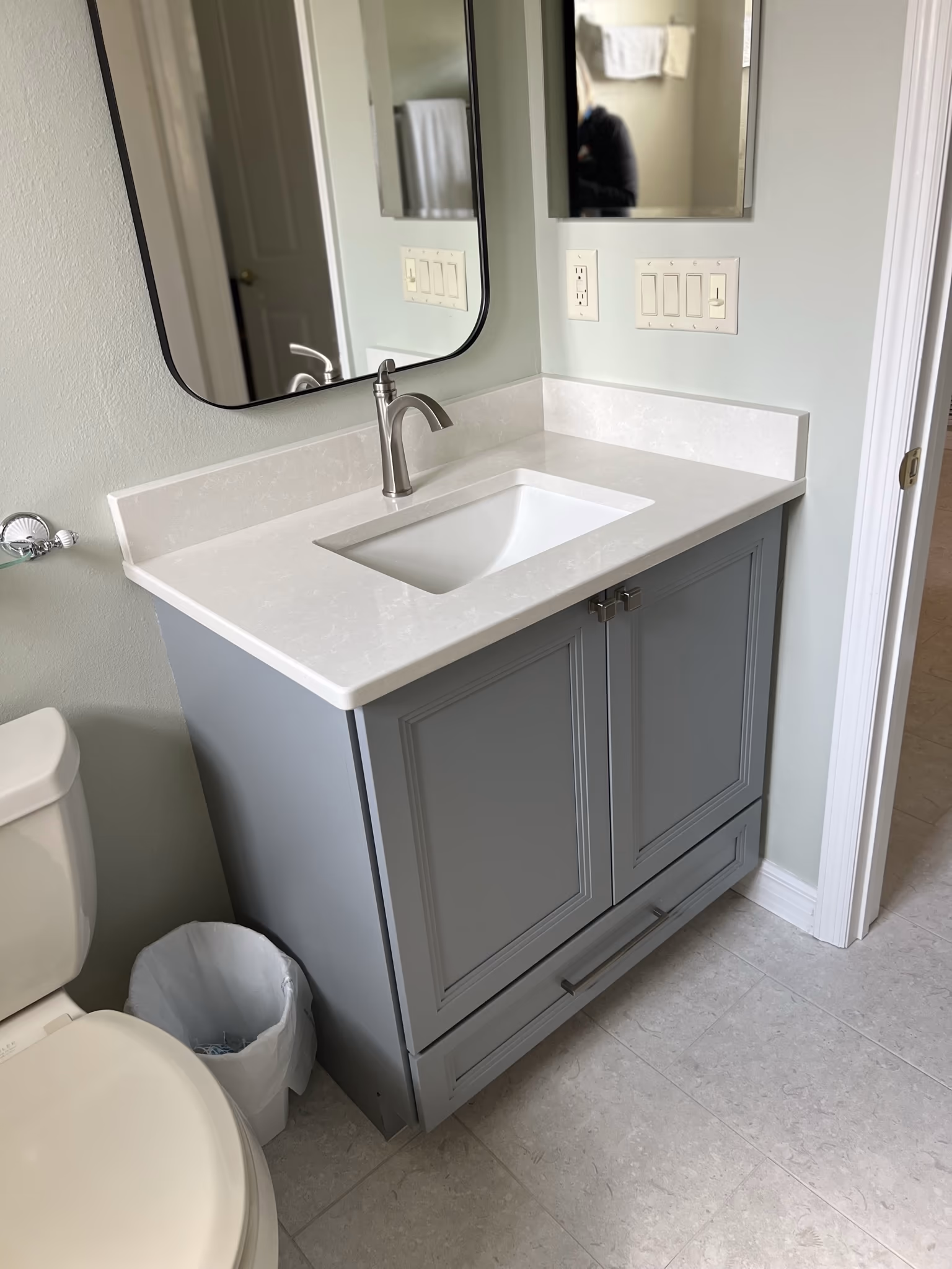Single-sink grey vanity with a white quartz top and black mirror, toilet and trash can visible at left