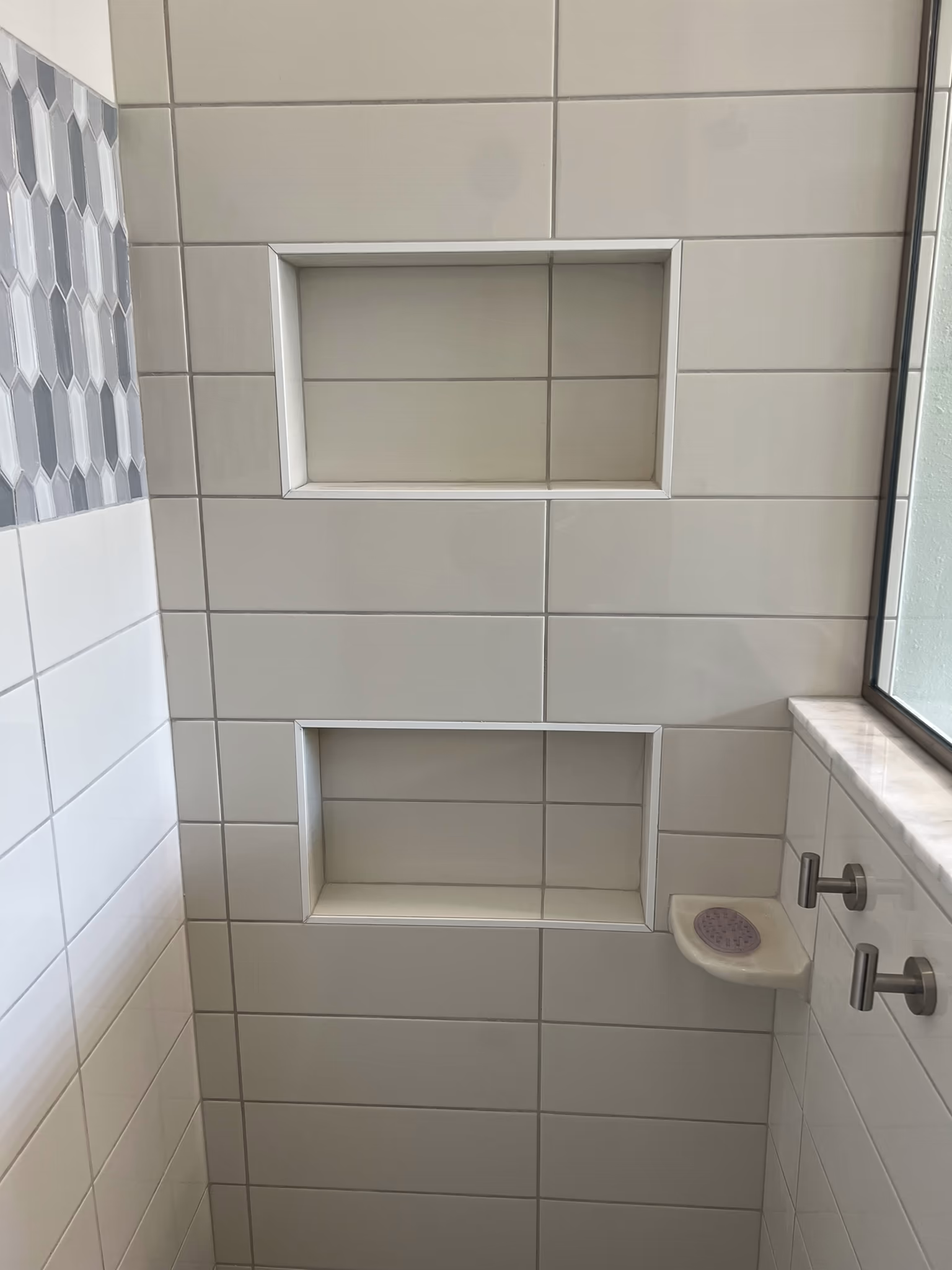 Two stacked tiled niches in a white subway tile shower wall, beside a marble ledge with towel hooks