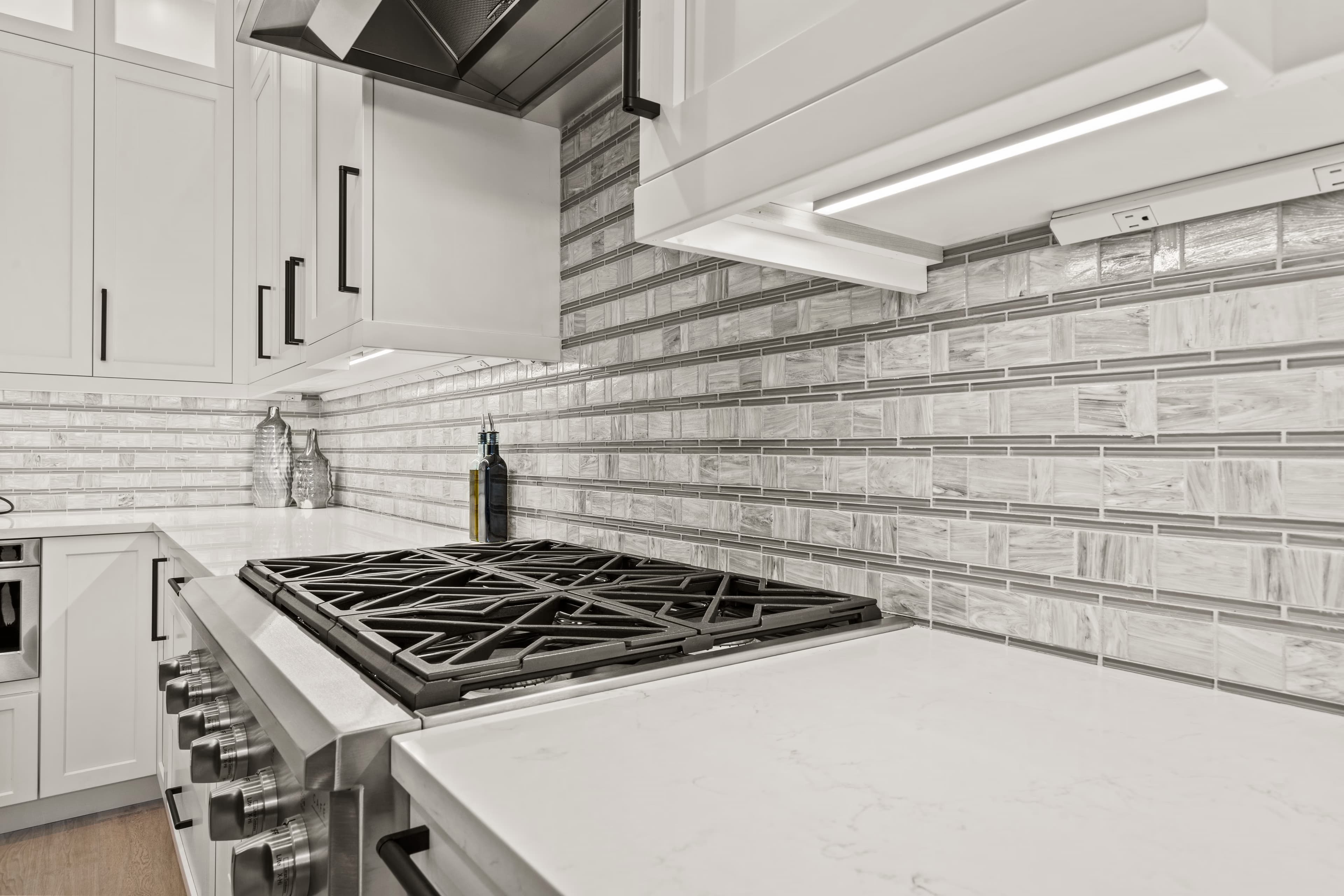 Close-up of a six-burner gas cooktop with a linear mosaic tile backsplash and quartz counter beneath the range hood