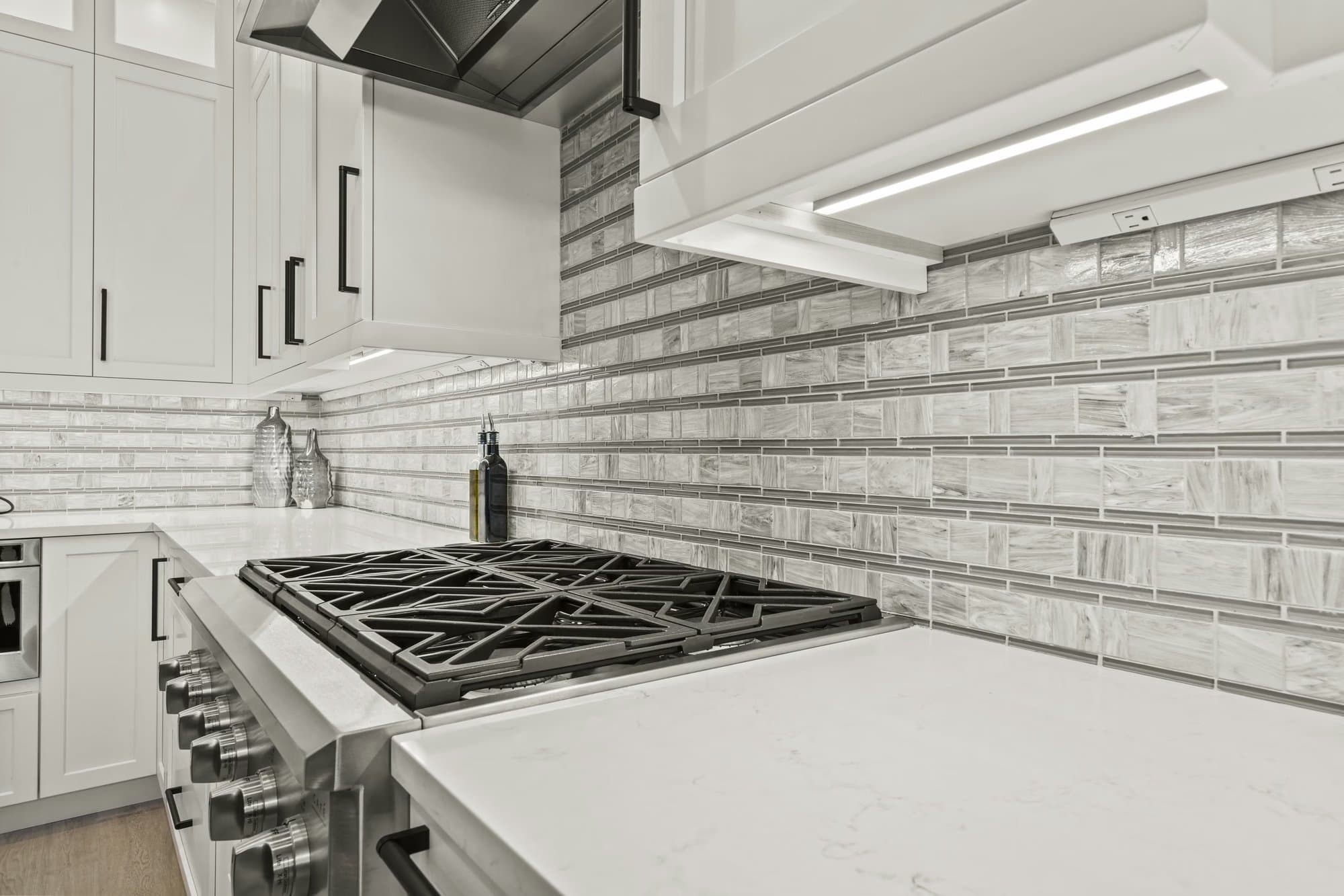 Close-up of a six-burner gas cooktop with a linear mosaic tile backsplash and quartz counter beneath the range hood