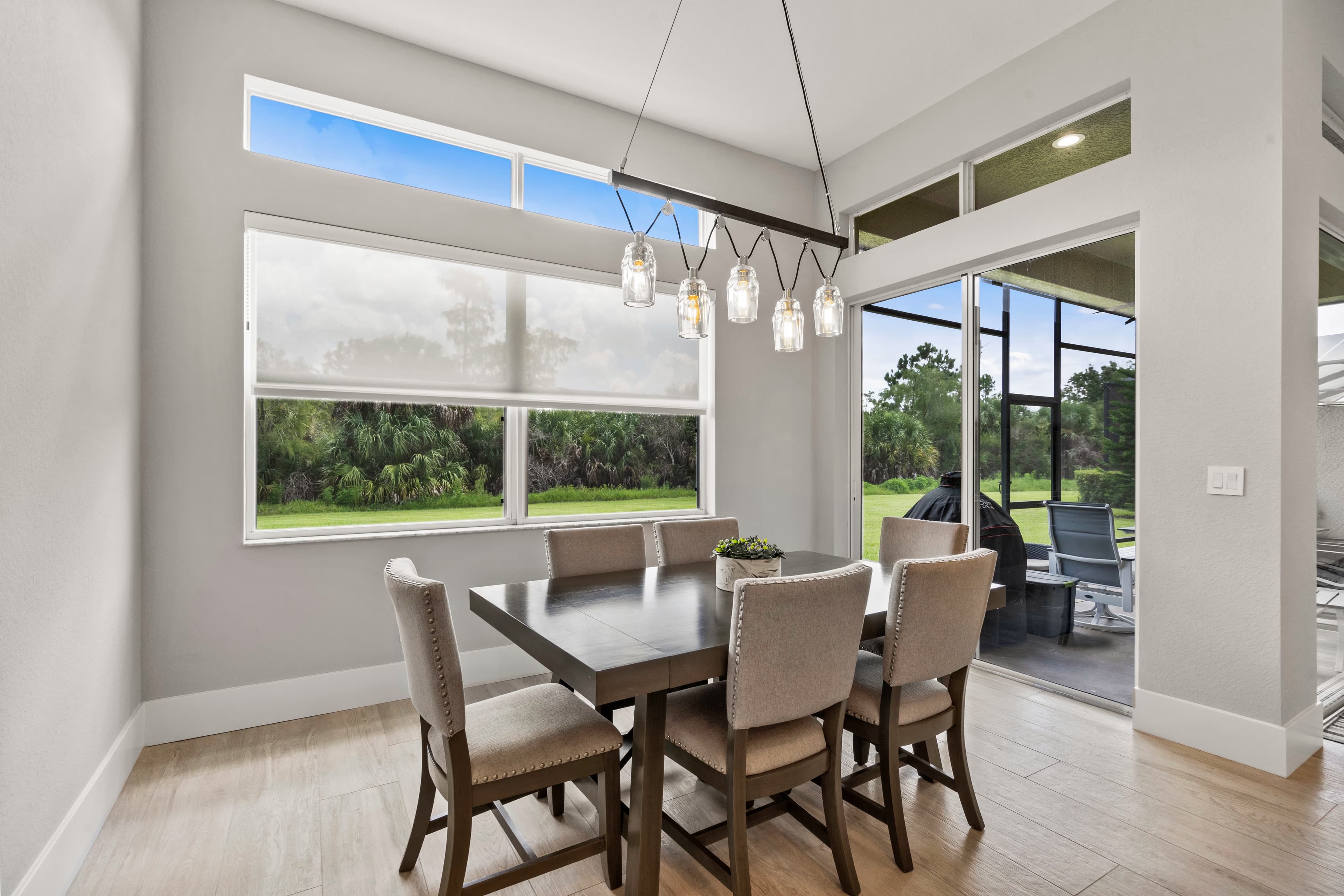 Dining table with six upholstered chairs under a pendant light, sliding glass doors open to a screened lanai with a grill