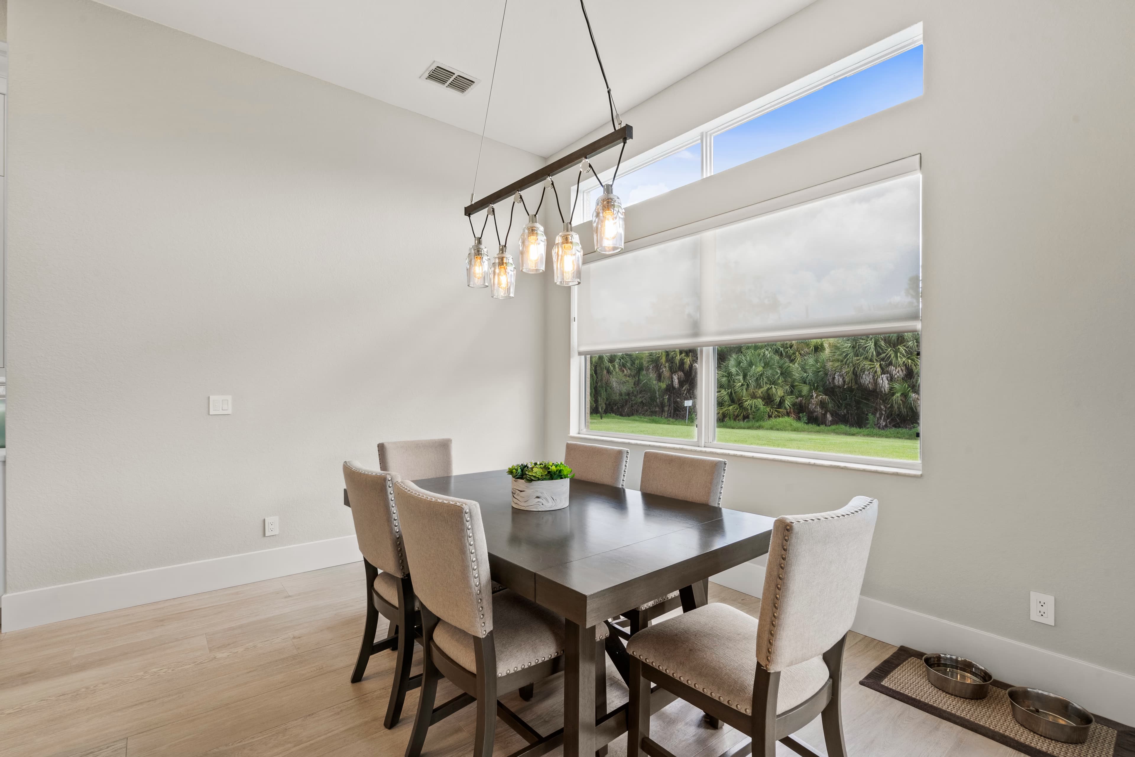 Dining table and chairs facing a large window with a sheer shade, pet food and water bowls on a mat near the floor