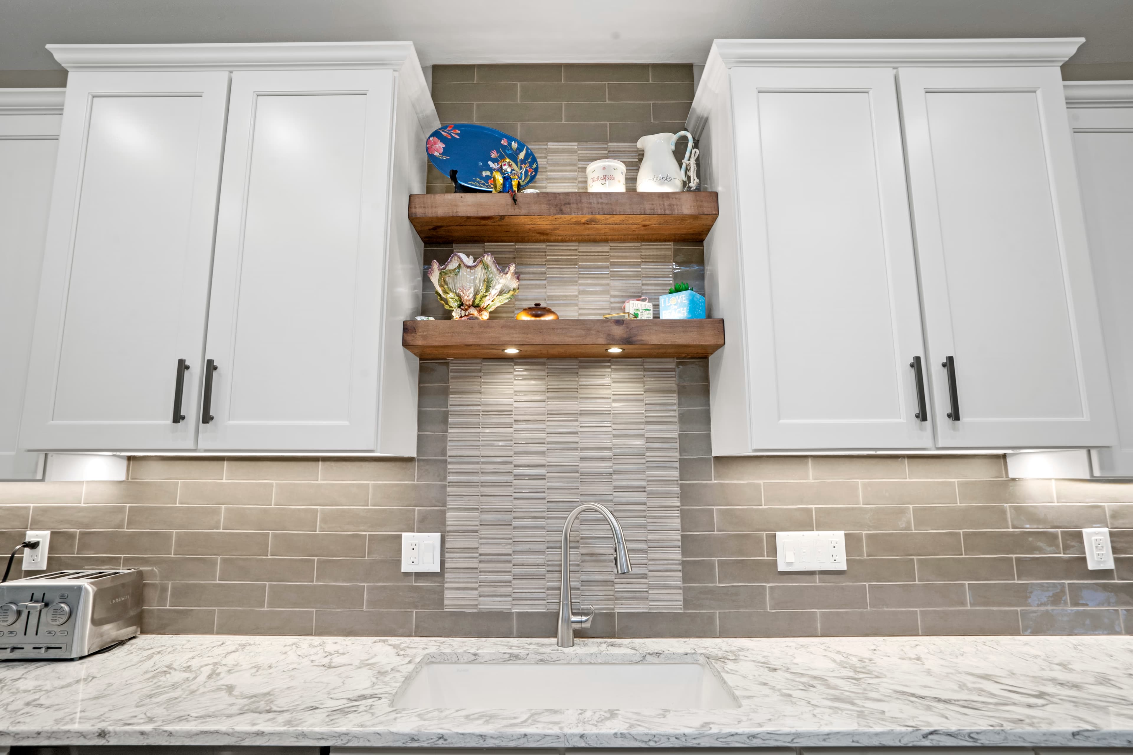 Wood floating shelves with a decorative pitcher and coastal decor above a marble-look counter and faucet