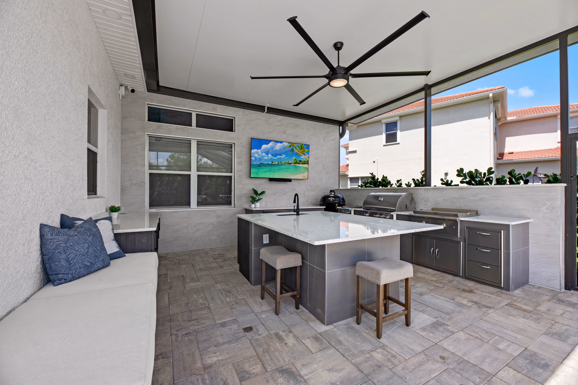 Covered lanai outdoor kitchen with a cushioned bench, ceiling fan, wall-mounted TV, and a quartz island with two stools