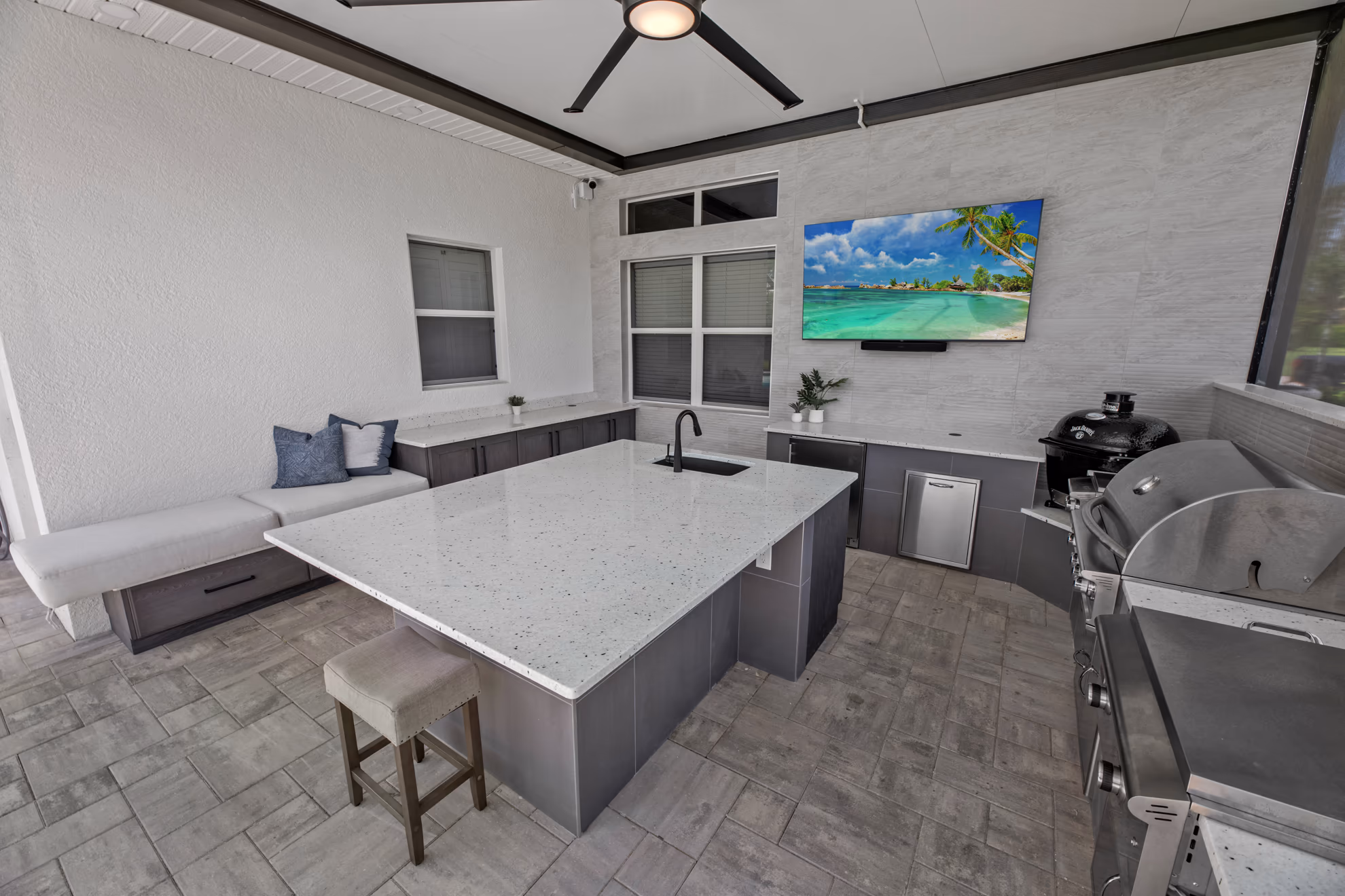 Outdoor kitchen seen from the bench, with a quartz island, wall-mounted TV, and built-in grill along the tiled wall