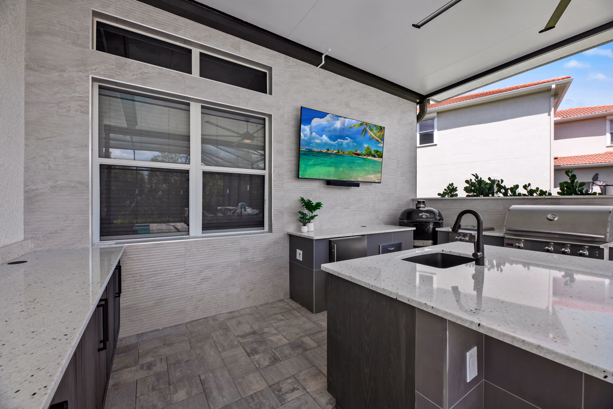 Outdoor kitchen sink and island beneath a wall-mounted TV, kamado grill to the right and a window with blinds on the left