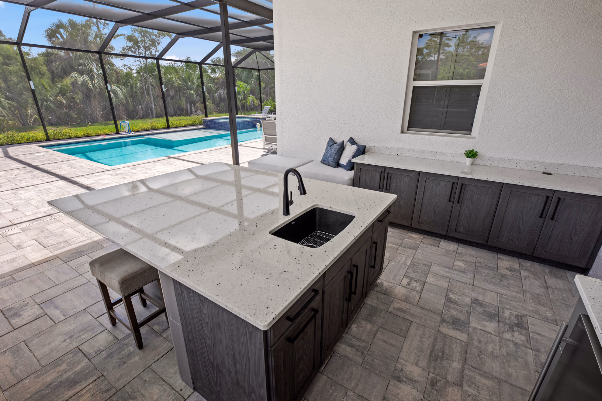 Outdoor kitchen island with a quartz counter and sink, next to a cushioned bench, overlooking a screened pool