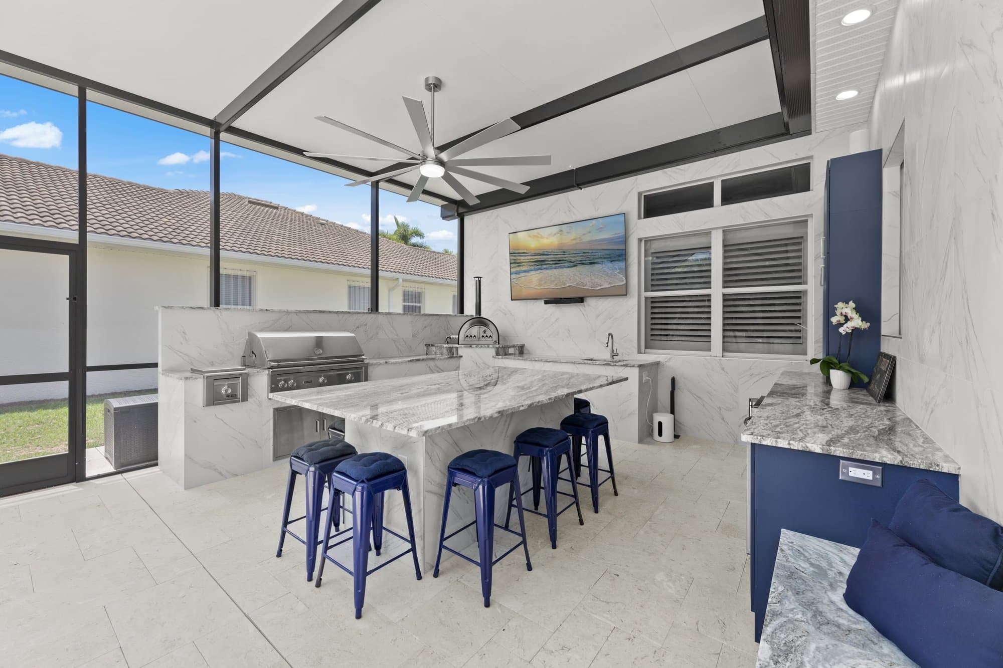 Screened lanai outdoor kitchen with a built-in grill and pizza oven, granite island with five navy stools, and navy cabinetry