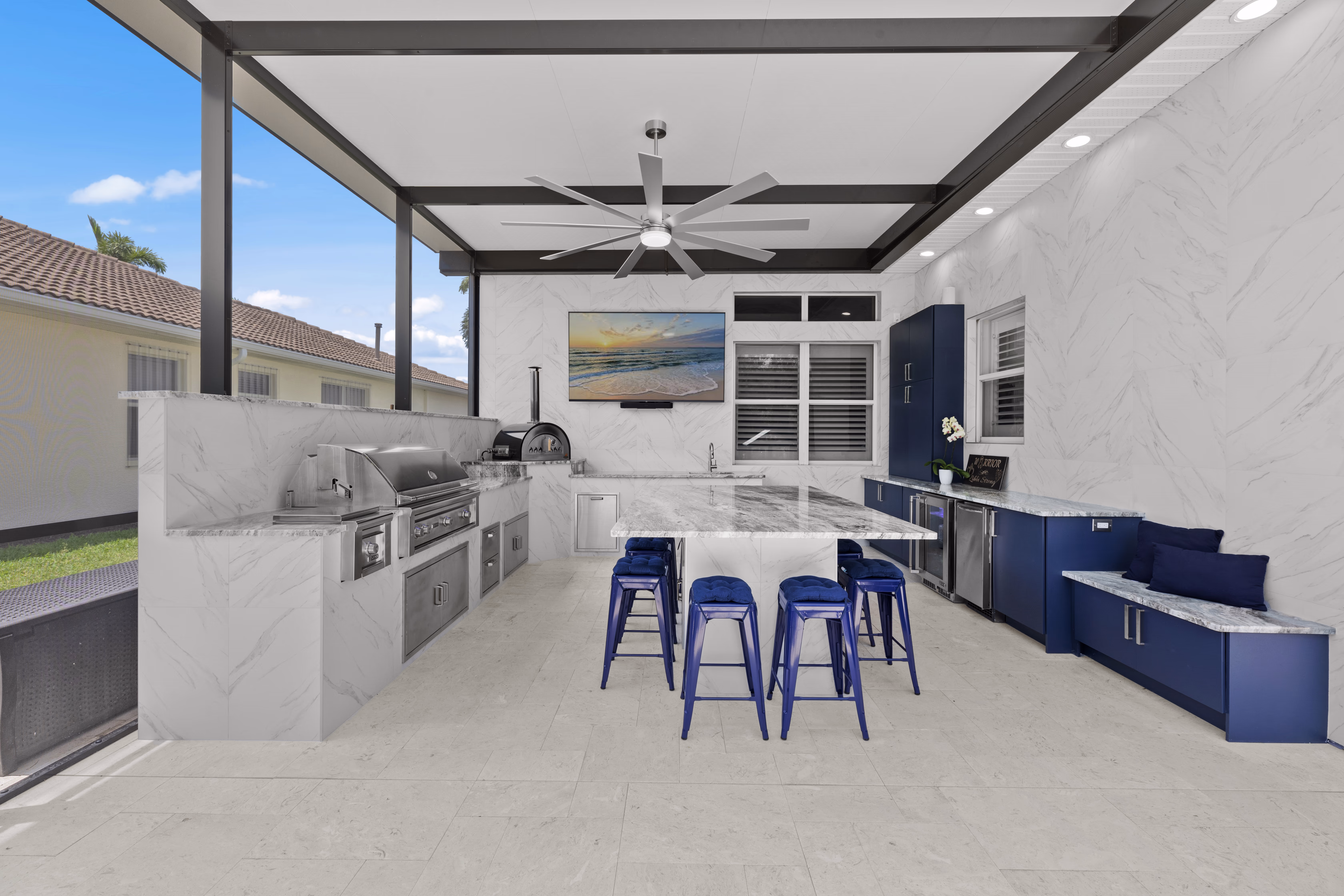 Wide view of an L-shaped outdoor kitchen with a grill, pizza oven, granite island, and navy cabinetry