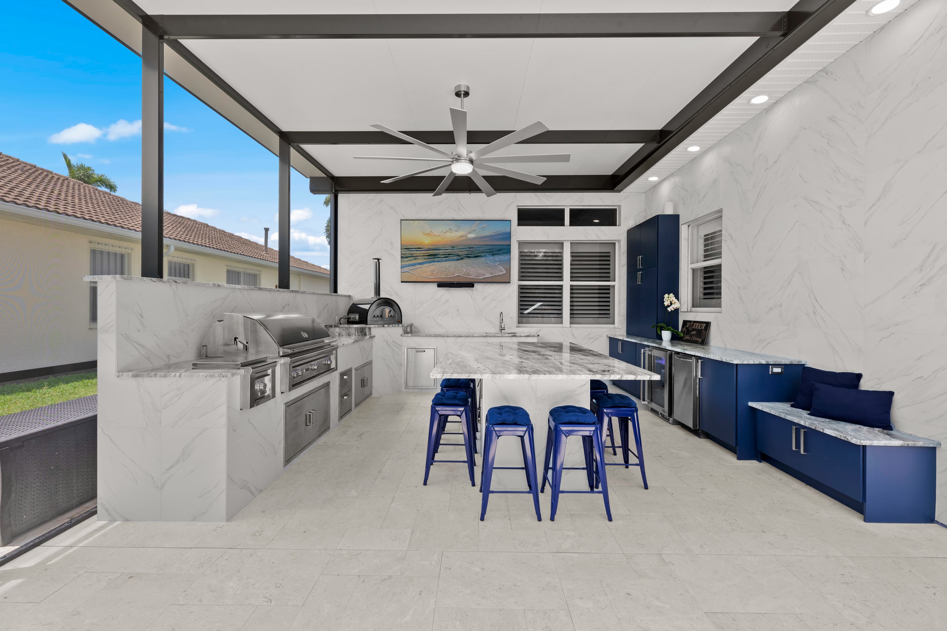 Wide view of an L-shaped outdoor kitchen with a grill, pizza oven, granite island, and navy cabinetry