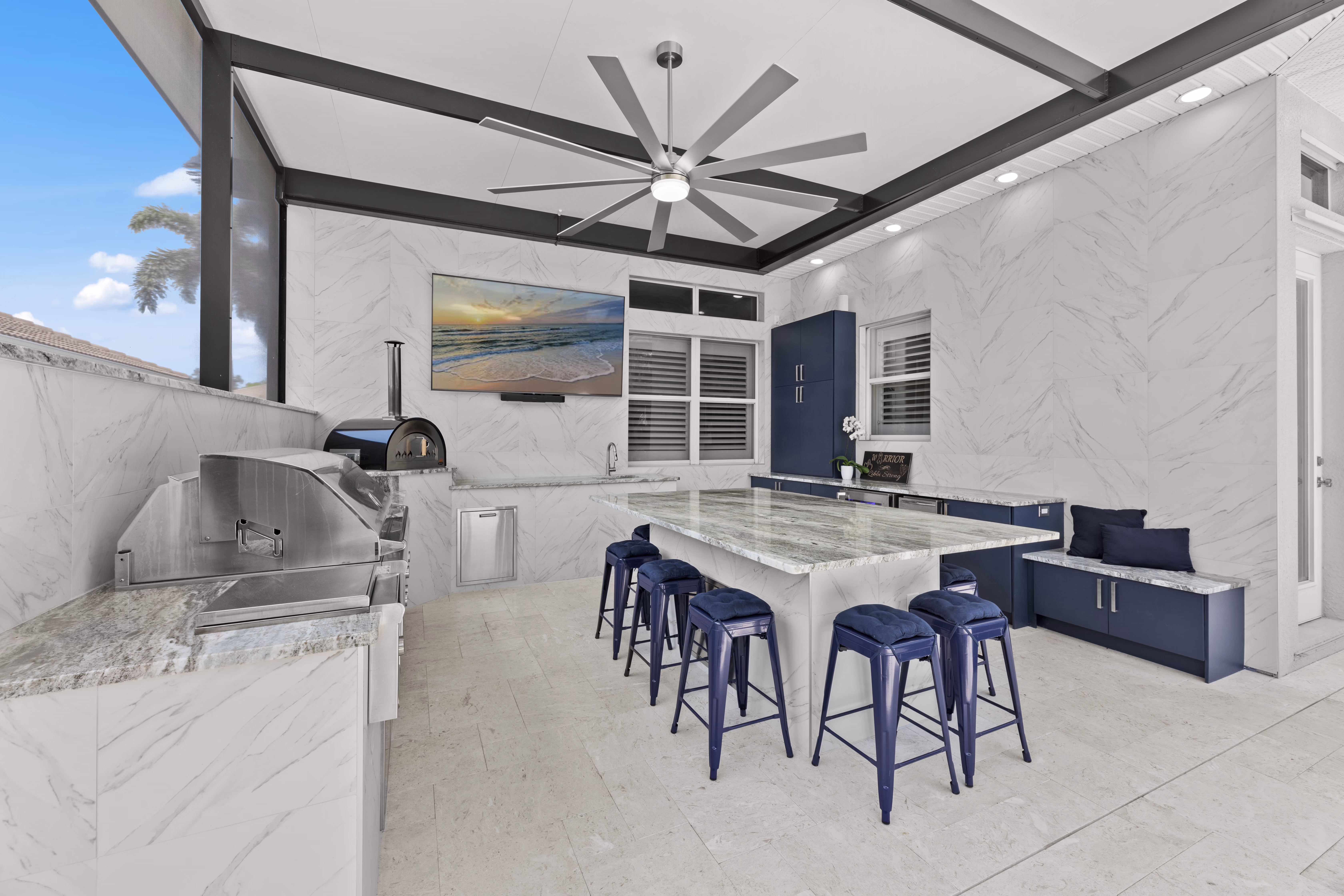 Outdoor kitchen with a pizza oven, built-in grill, granite island with navy stools, and a bench with navy pillows