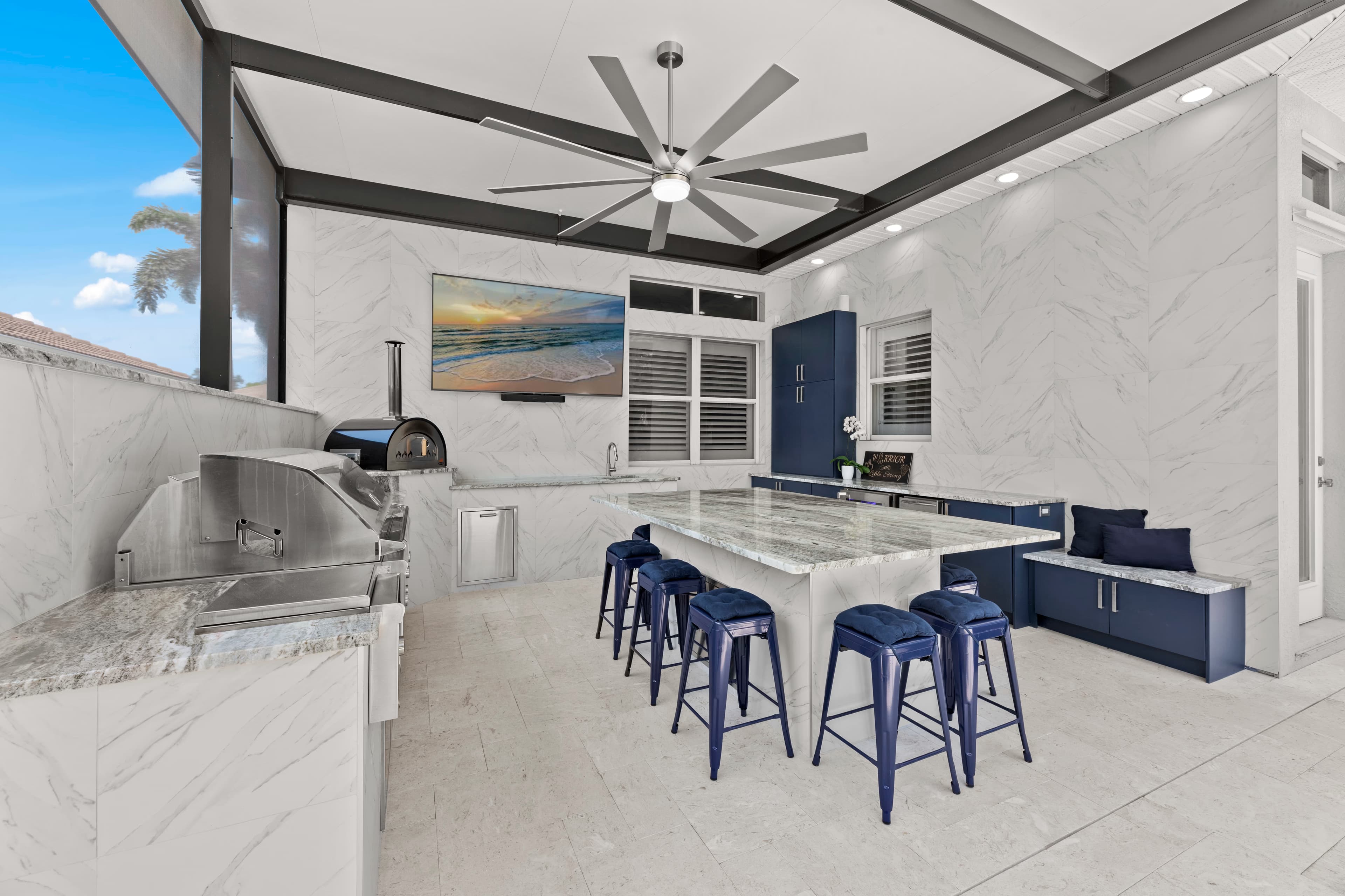 Outdoor kitchen with a pizza oven, built-in grill, granite island with navy stools, and a bench with navy pillows