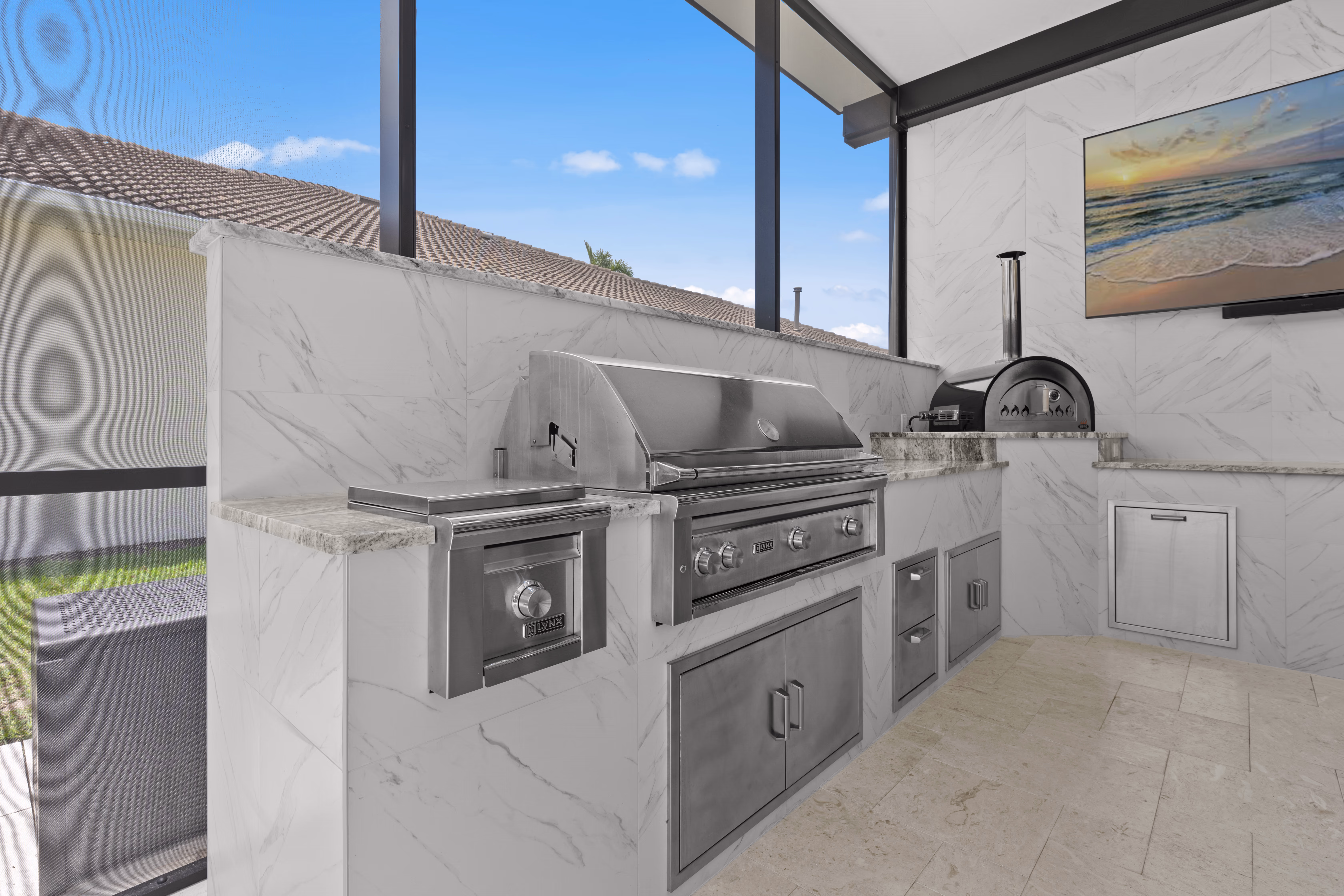 Stainless grill station with a warming drawer and storage doors, marble counter, and a pizza oven beyond