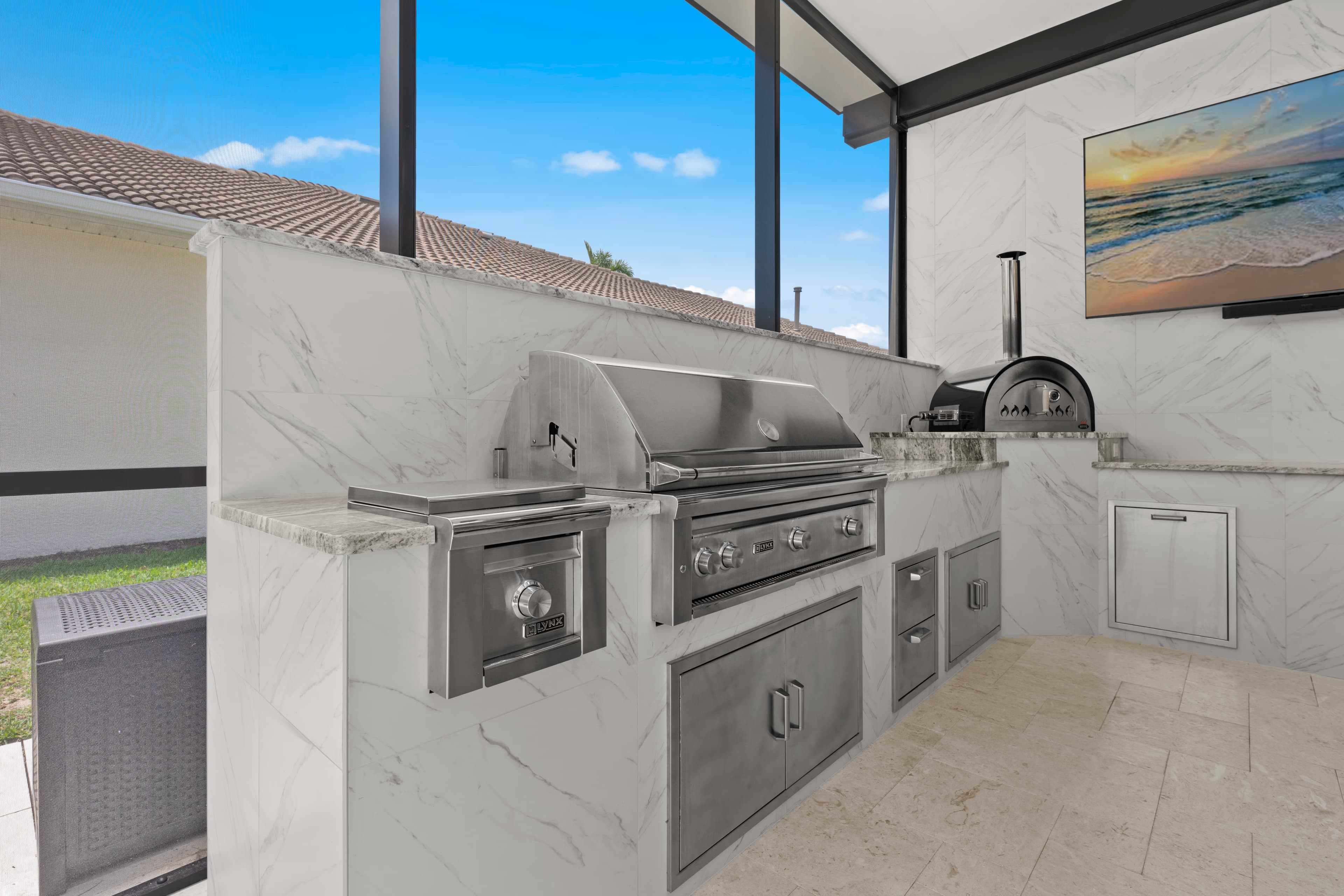Stainless grill station with a warming drawer and storage doors, marble counter, and a pizza oven beyond