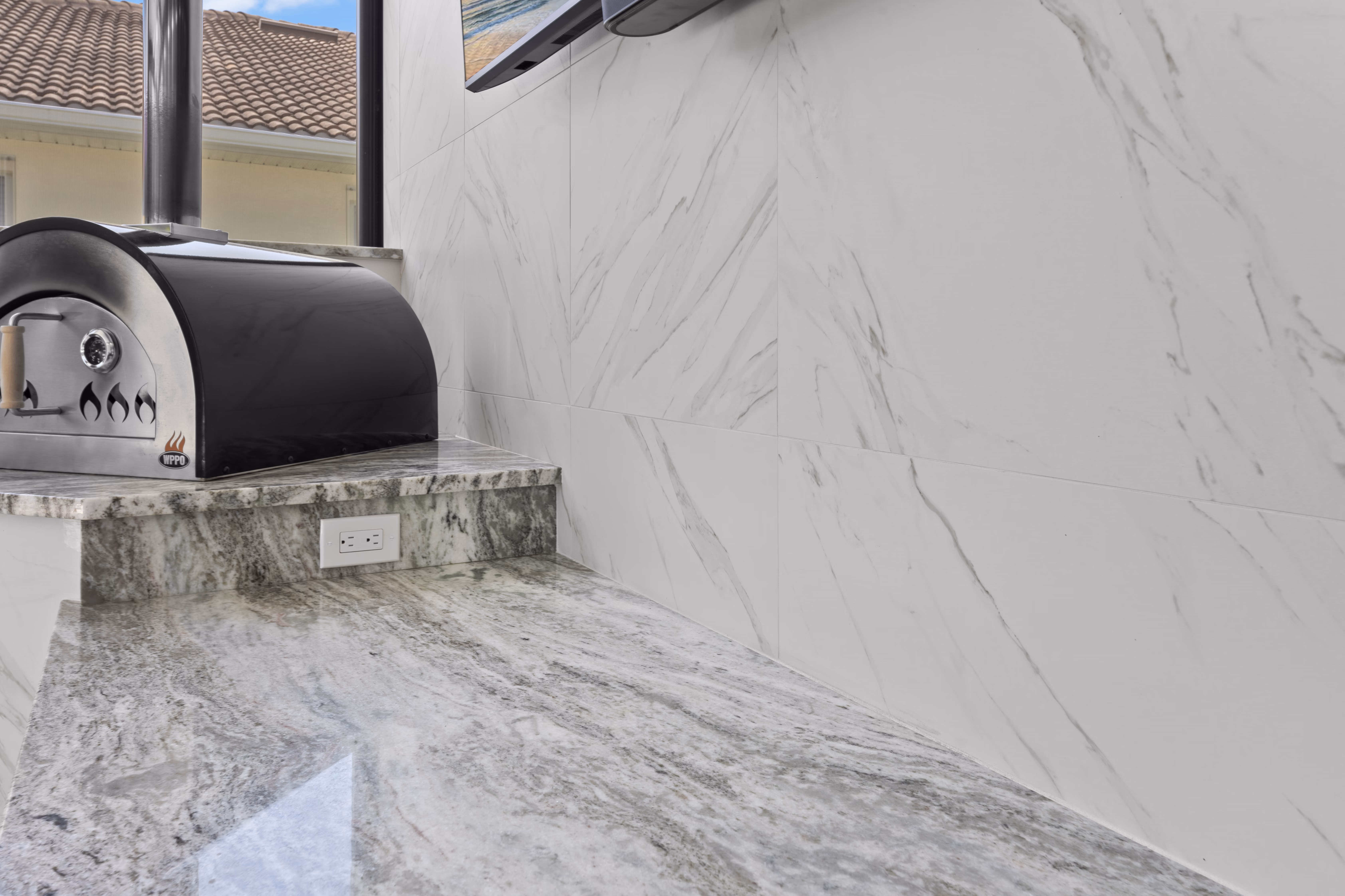 Close-up of a black wood-fired pizza oven on a marble ledge beneath a wall-mounted TV