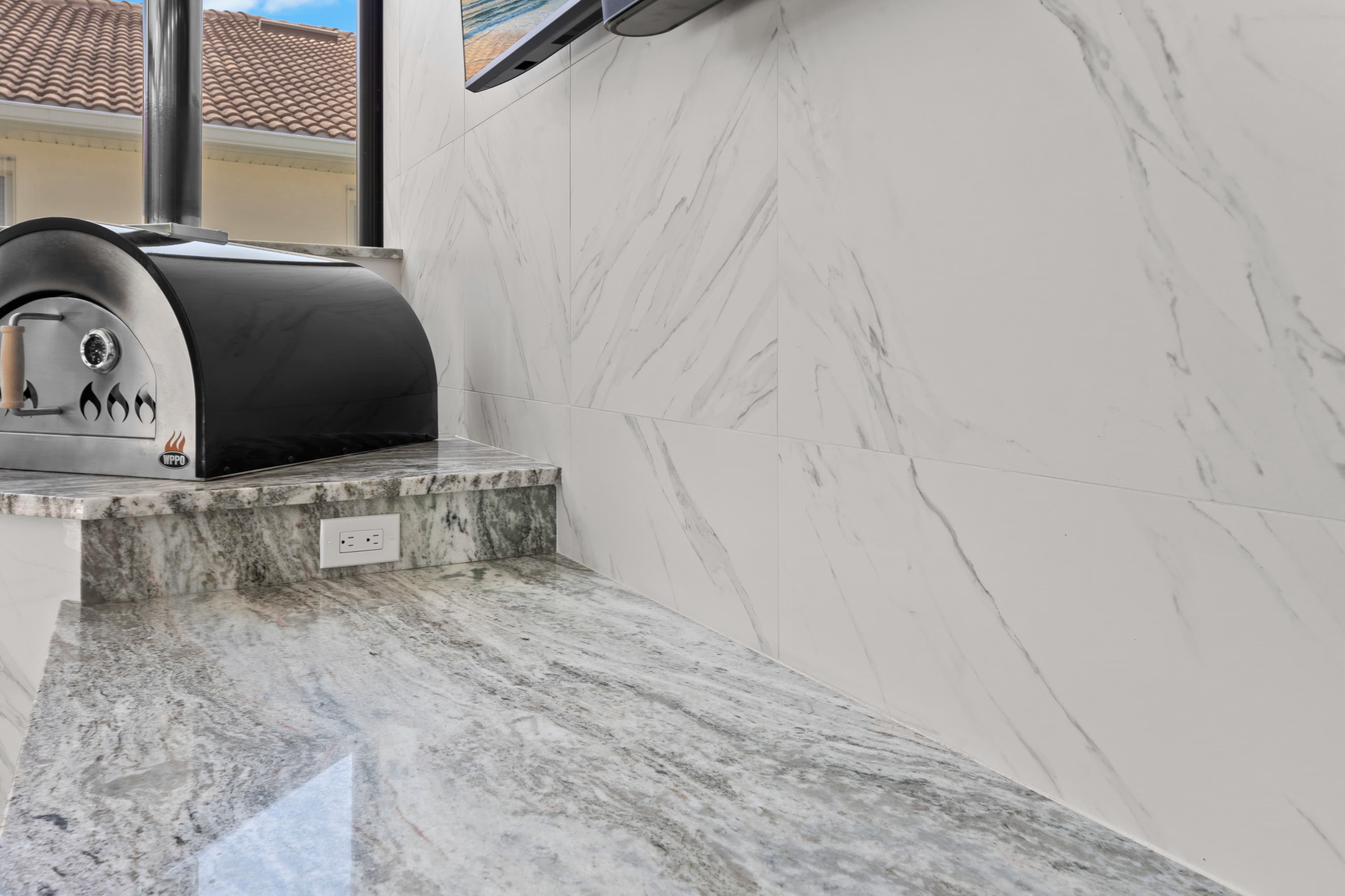 Close-up of a black wood-fired pizza oven on a marble ledge beneath a wall-mounted TV