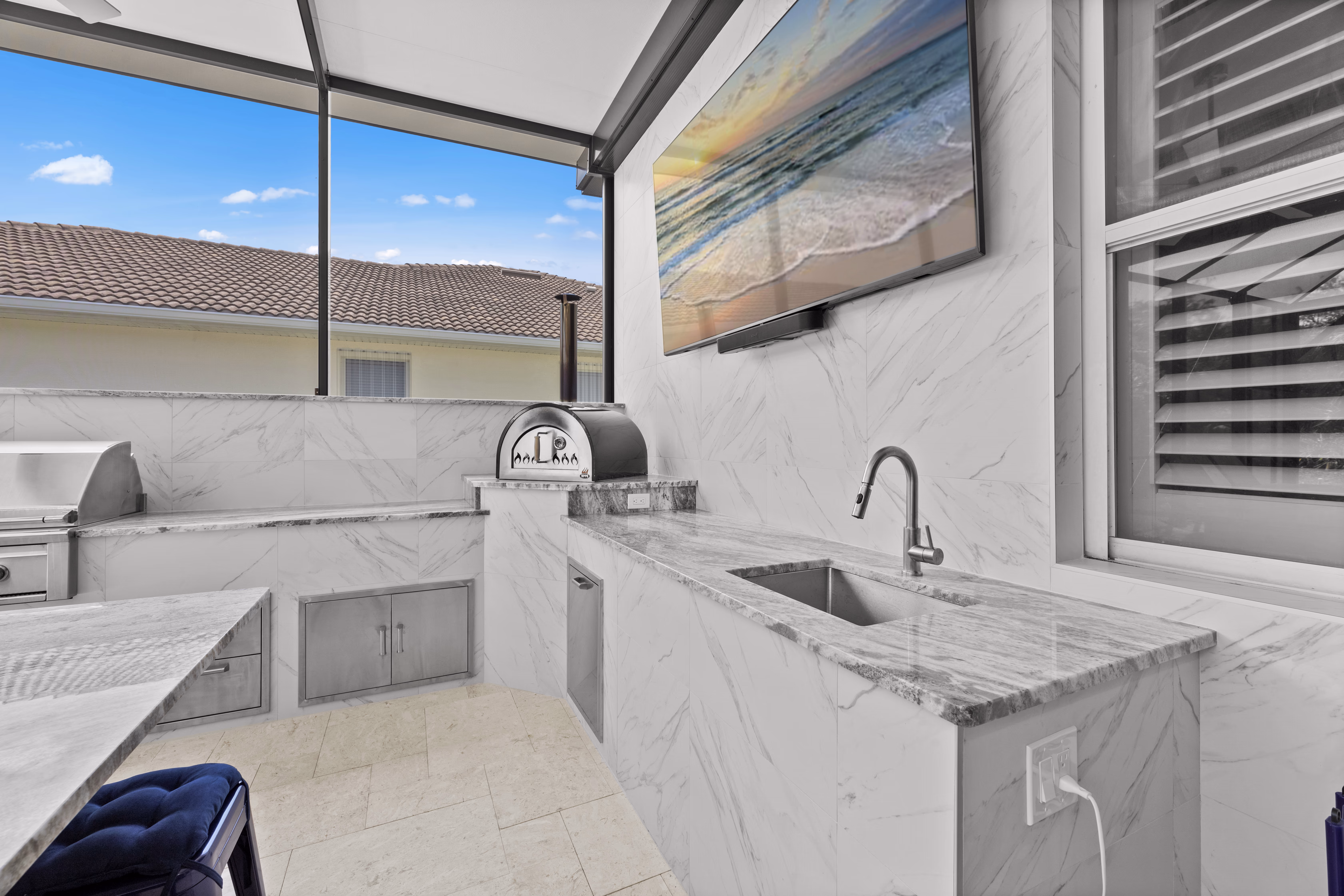 Pizza oven and a sink with a gooseneck faucet on a marble counter, beneath a wall-mounted TV
