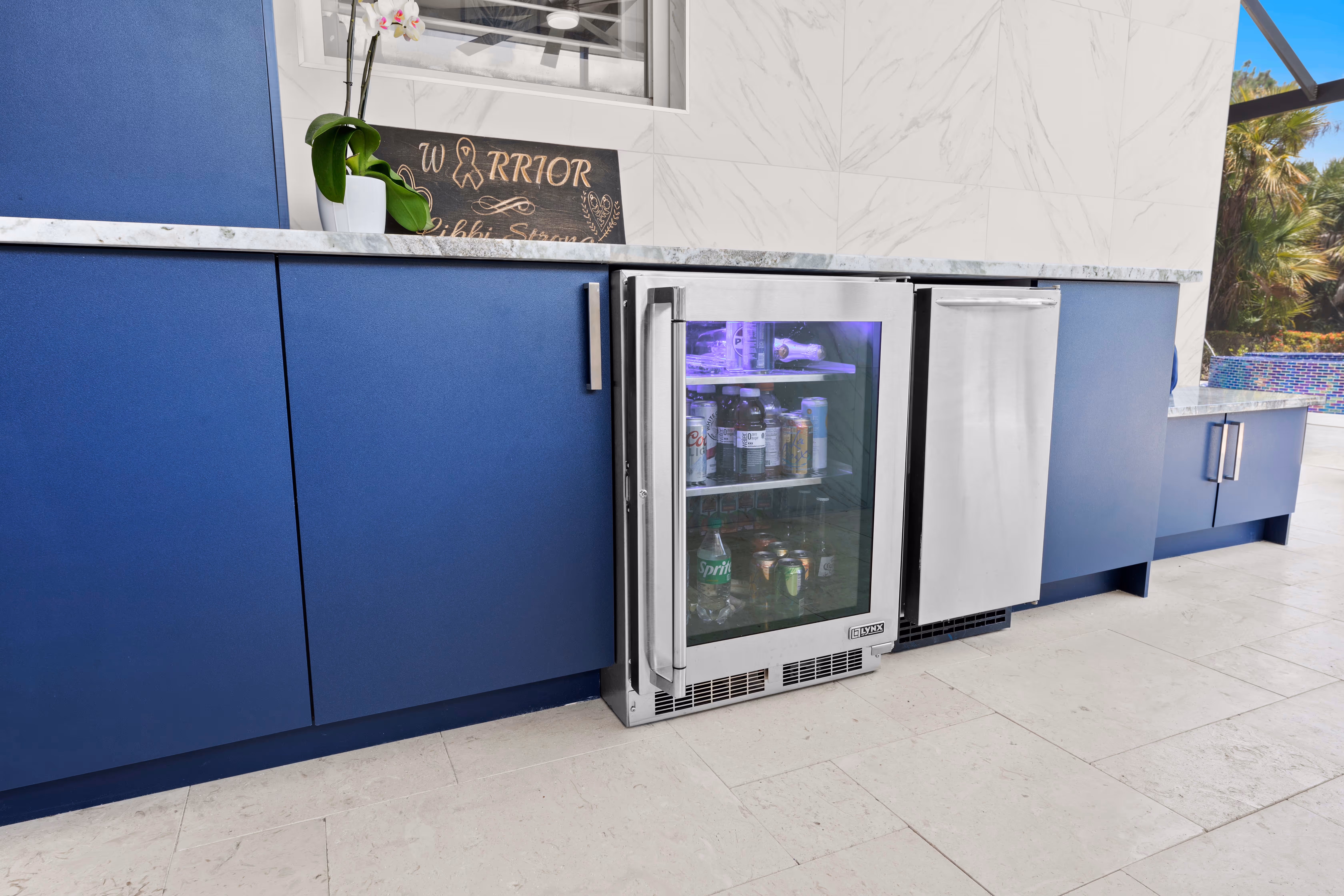 Close-up of a stocked glass-door wine cooler and stainless mini fridge built into navy outdoor cabinetry