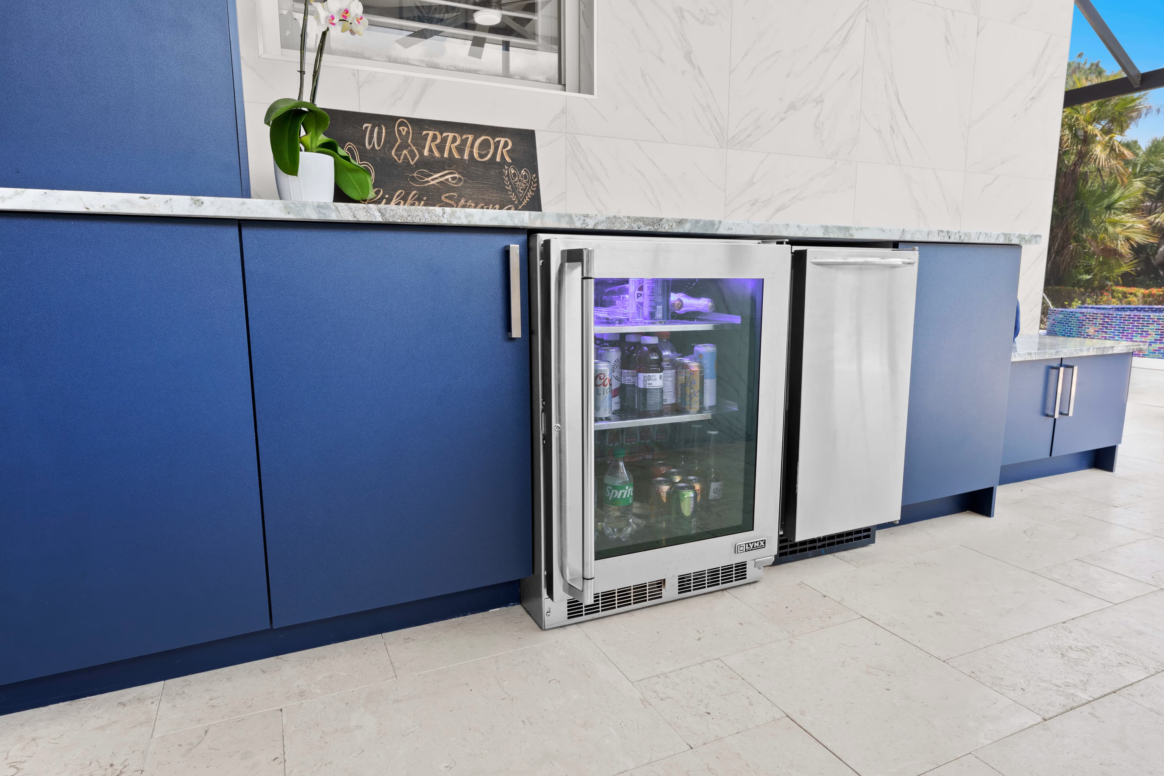 Close-up of a stocked glass-door wine cooler and stainless mini fridge built into navy outdoor cabinetry